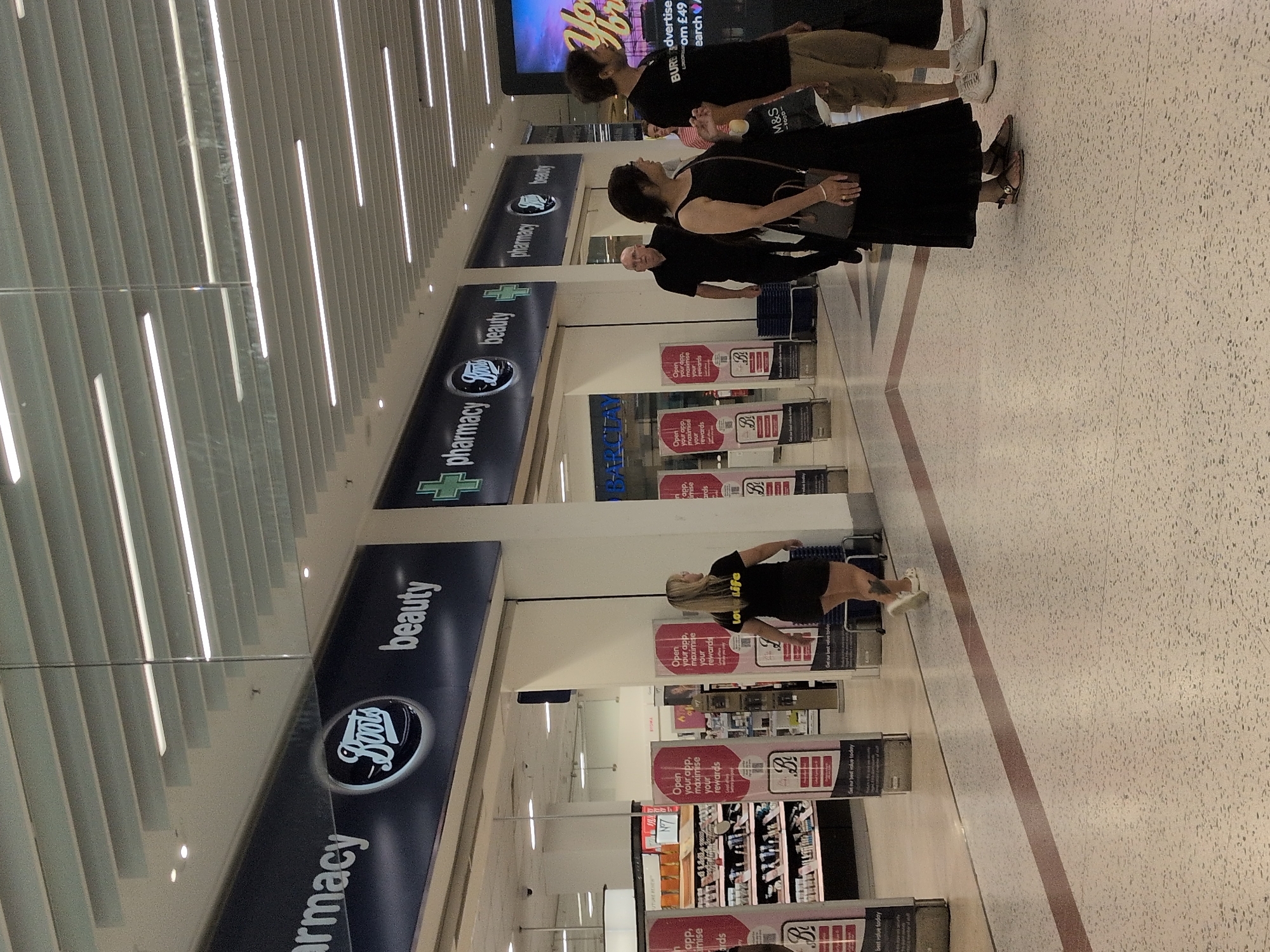 The image depicts an indoor shopping center, likely a Boots pharmacy and beauty store, given the prominent signage. Several individuals are visible walking through the space. On the left, a woman with blonde hair is walking away from the camera, wearing a black t-shirt with "love life" printed on it and black shorts. To her right, a bald man in a black shirt is walking towards the viewer. Further to the right, a woman in a black dress with a handbag is walking away, and a man in a black t-shirt and khaki shorts is also visible. The environment features illuminated ceiling panels and floor-to-ceiling glass partitions. Promotional posters are displayed throughout the store. The flooring is a terrazzo-style material with a red stripe demarcating a walkway. The overall impression is a busy retail setting within a modern shopping mall.