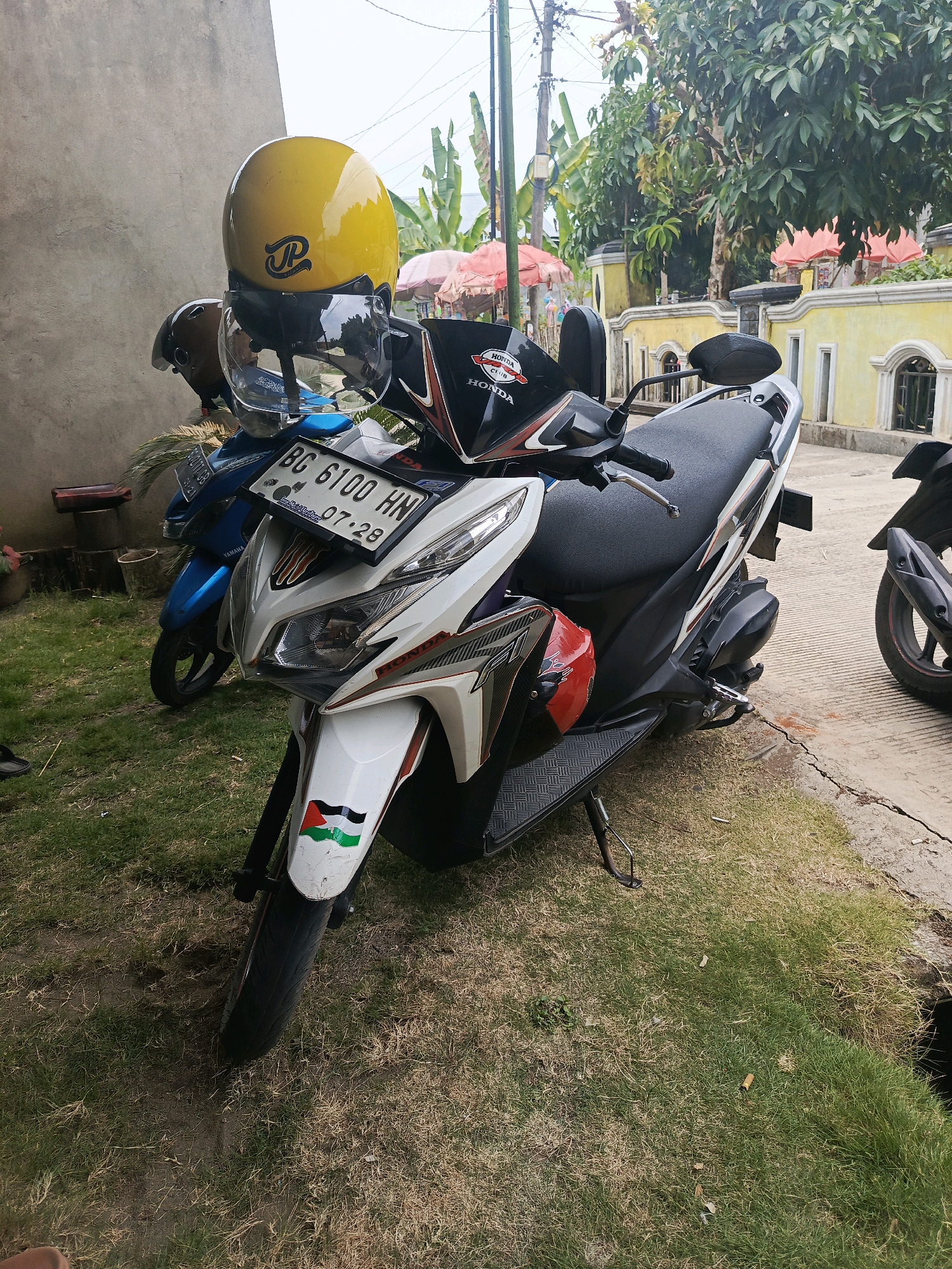 Two scooters are parked on a grassy area next to a concrete path. The scooter in the foreground is white with red and black accents and has a license plate that reads "BG 6100 HN 07-28". A sticker of the Palestinian flag is visible on the front of this scooter. A yellow helmet with a clear visor sits on the handlebars of this scooter, and a brown helmet is behind it. A blue scooter is parked behind the white one, with its license plate partially visible. In the background, there is a yellow building with decorative windows and a stucco-like wall. Several trees and some pink umbrellas are also visible in the background. The scene appears to be outdoors in a residential or public area in Lubuklinggau, Indonesia.