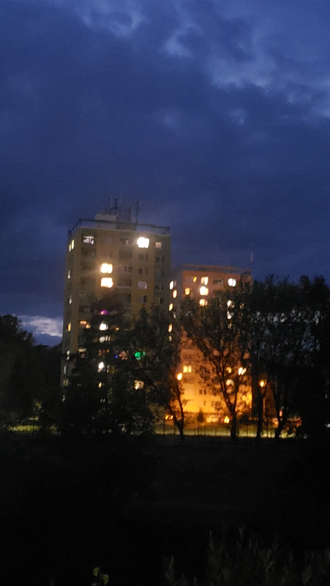 The image depicts two apartment buildings at dusk, illuminated by interior lights. The larger, yellow building on the left is taller than the orange building on the right. Both structures are residential, with multiple windows visible, many of which are lit. Trees and foliage are present in the foreground, obscuring the lower portions of the buildings and the ground. The sky above is a deep blue, with some cloud cover. The scene appears to be an urban environment, likely a residential area within the city of Prešov, Slovakia, given the architectural style. There are no people visible in the image. The overall impression is one of nighttime in a city.