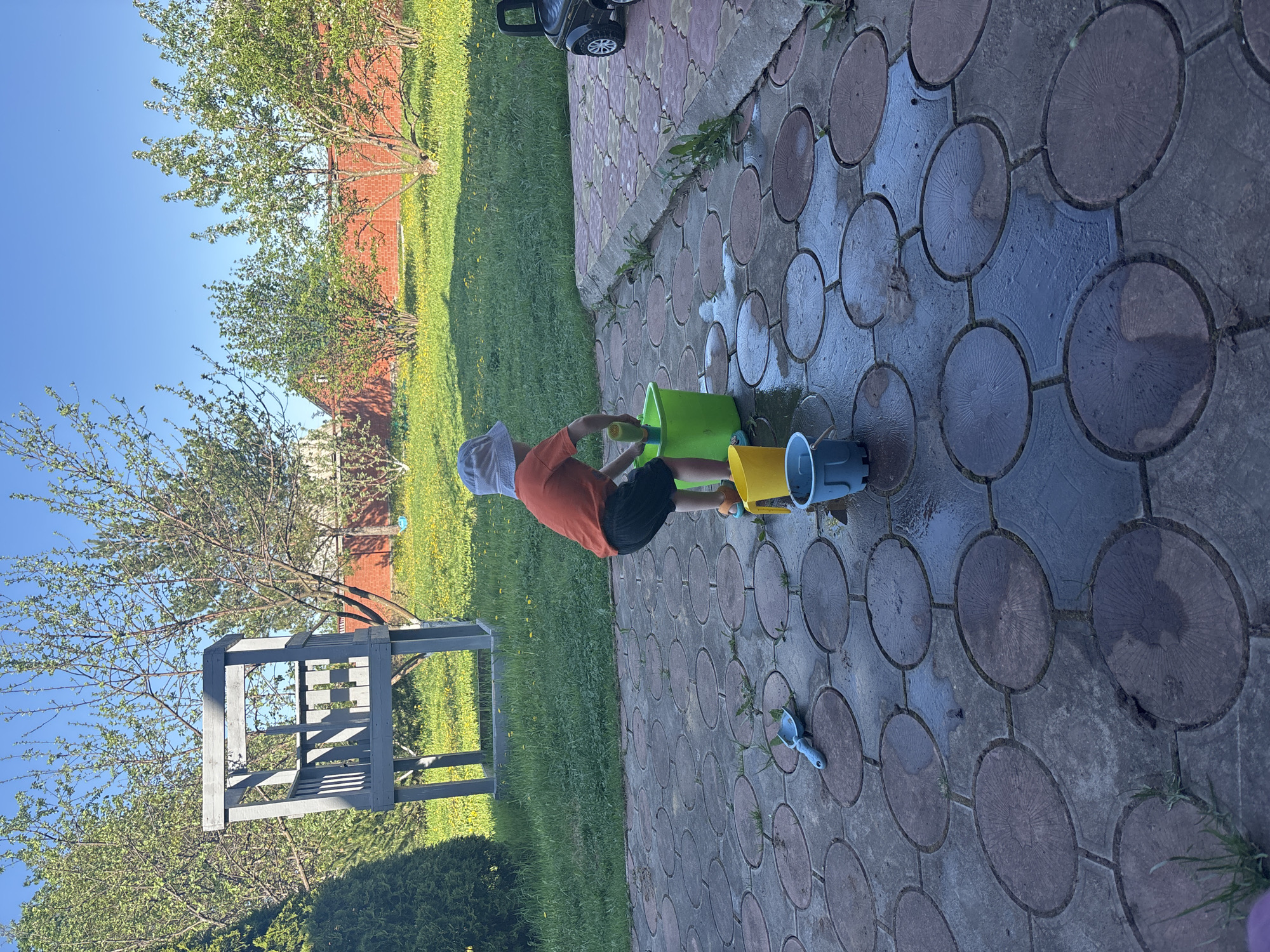 A child in an orange shirt and white hat is playing with water and toys on a paved area of circular stones. A green bucket, yellow bucket, blue bucket, and a small blue shovel are near the child. The paved area is adjacent to a grassy area with a wooden structure and trees. In the background is a red brick building. The location is Ферма, Russia.