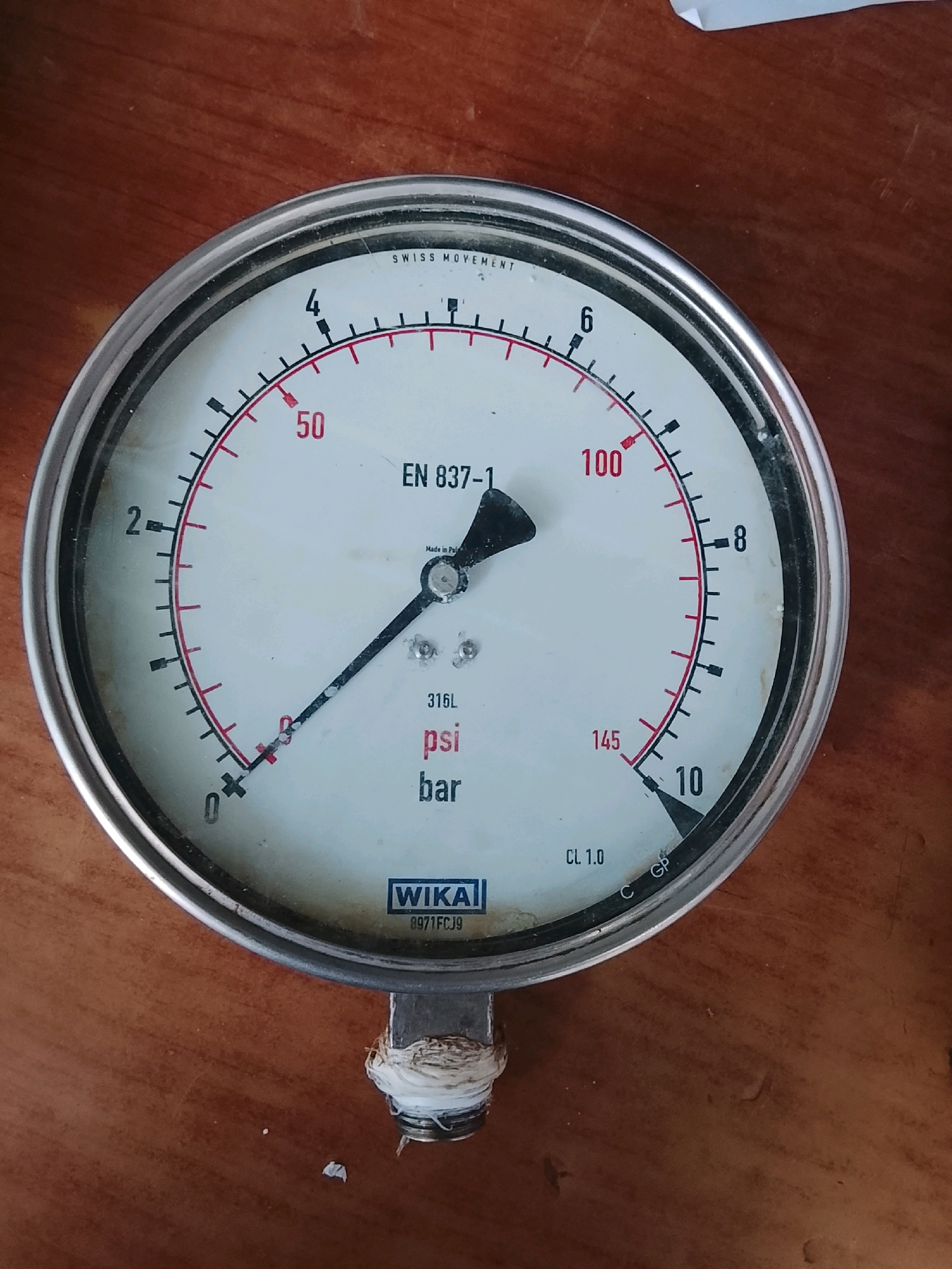 The image shows a close-up, top-down view of a pressure gauge resting on a wooden surface. The gauge is round with a silver metal rim and a white face. It has black markings and red accents indicating pressure measurements. A black needle points to a value slightly above 20 on the scale. The scale ranges from 0 to 10, and also shows markings for 50, 100, and 145, with corresponding units labeled as "psi" and "bar". Text on the gauge includes "SWISS MOVEMENT", "EN 837-1", "316L", "WIKA", and "8971FCJ9". The wooden surface appears to be a table or desk. There is no indication of the city of Pataar Goth, Pakistan, or any other specific location in the image.