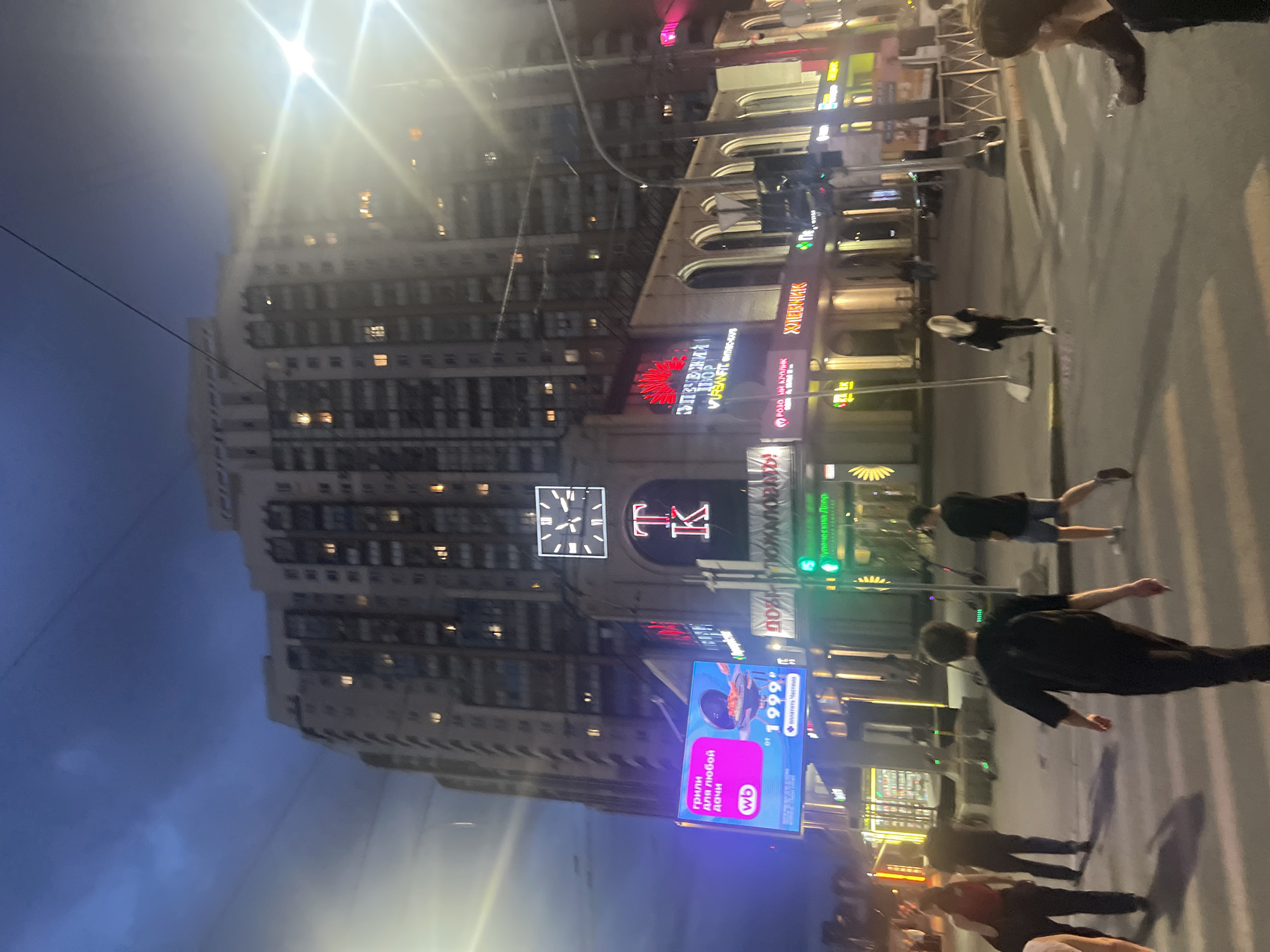 This nighttime urban photograph depicts a bustling street scene in Saint Petersburg, Russia, under an overcast, dark blue sky. A tall, multi-story building, likely residential or mixed-use, dominates the background, with numerous illuminated windows. On its facade, a large square digital clock with white Roman numerals, indicating approximately 11:50 or 12:50, is positioned above a prominent red glowing "TK" sign. The building's lower levels house commercial establishments, featuring multiple illuminated signs with Cyrillic text in various colors. A large blue digital billboard on the left advertises "ГРИЛИ для любой дачи" (Grills for any dacha) with a Wildberries "wb" logo and a price of "от 1999 ₽". Other visible signs include "URBANFIT fitness-club" in red, "ХЛЕБНИК", and "Лучевой Двор" in green, alongside a green traffic light. The street level, marked by a faint zebra crossing, shows approximately seven to ten pedestrians. In the foreground, a man with a black backpack walks away from the camera, while another man in a dark t-shirt and shorts walks towards the right. A woman with long blonde hair, dressed in black, walks away further into the midground. Several electric scooters are parked along the curb. A bright streetlight on the right emits a strong flare, and overhead wires are visible against the sky.
