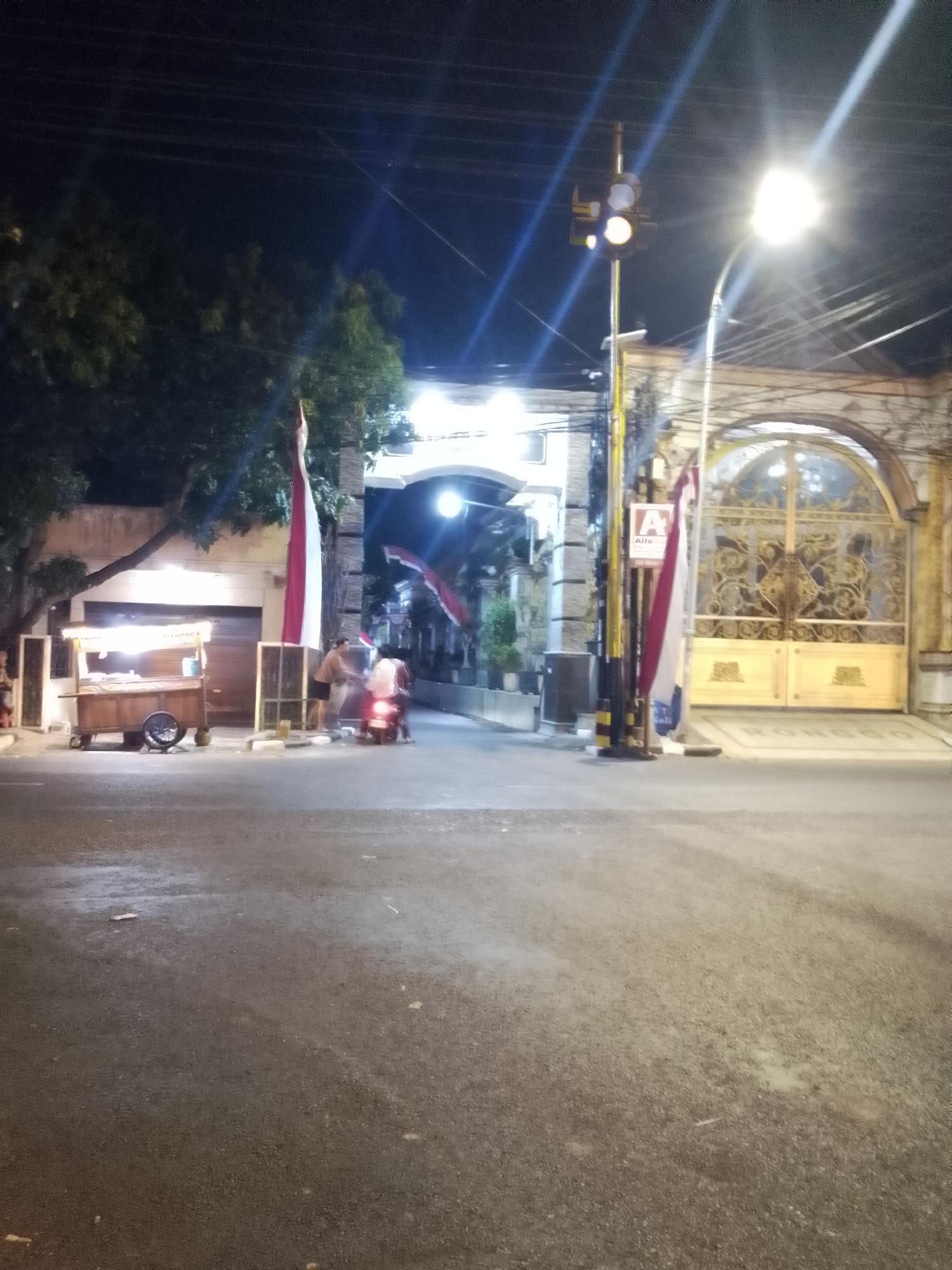 A nighttime street scene in Lamongan, Indonesia shows a food cart with a red and white Indonesian flag displayed on it, parked on the left side of the asphalt road. To the right of the food cart, two people are interacting near a motorcycle, one of whom is wearing a white shirt and appears to be handing something to the other. Further to the right, a large ornate golden gate is visible, flanked by pillars and decorative fencing. A traffic light with a yellow lamp illuminated is positioned near the gate. The street is illuminated by streetlights and the lights from the food cart. The environment appears to be an urban setting with trees and buildings visible in the background.