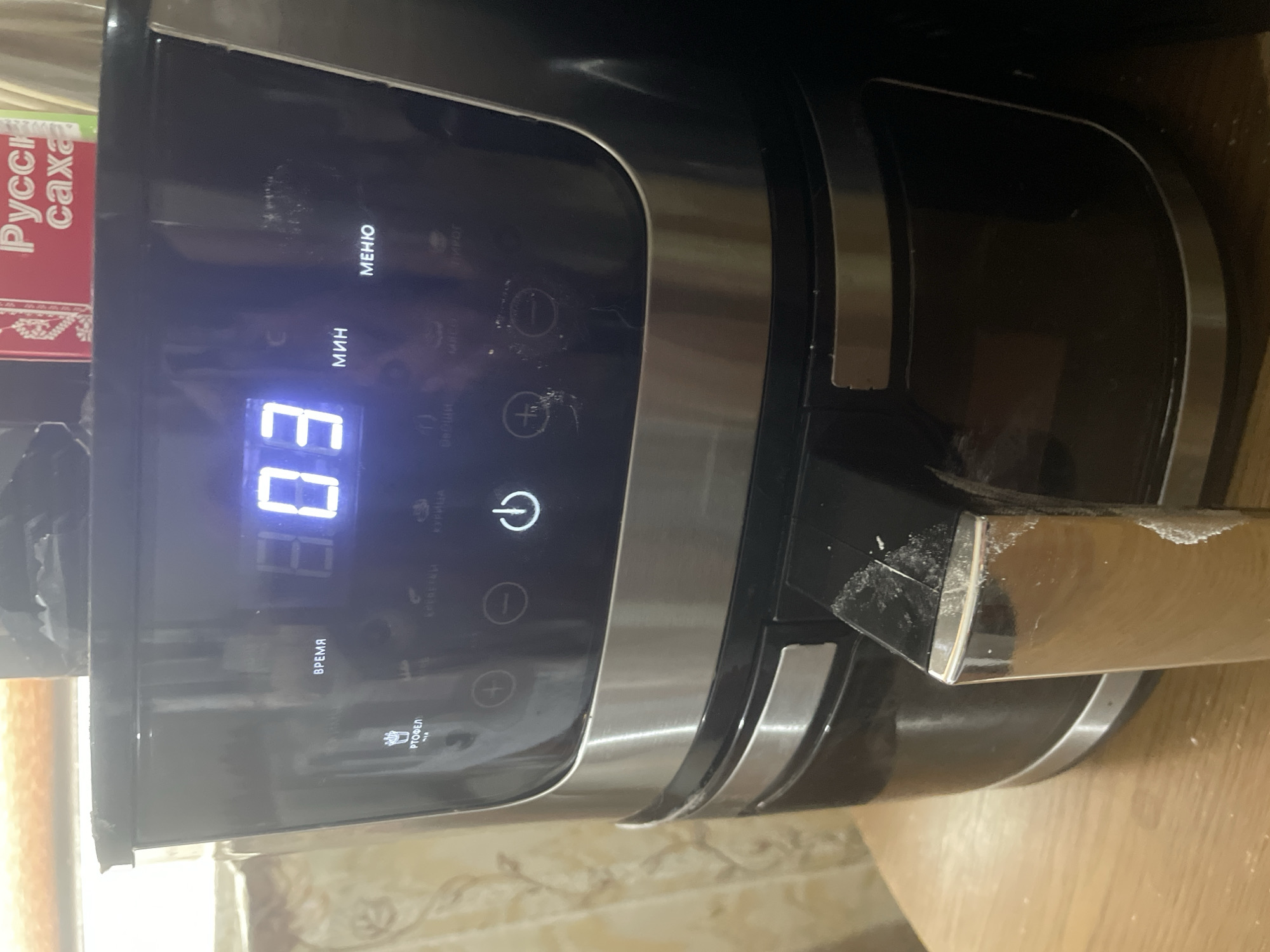 This media file shows a close-up of a black air fryer with a digital display. The display shows "03" in large, illuminated blue digits, likely indicating minutes remaining in a cooking cycle. Above the digits, the word "ВРЕМЯ" (TIME) is visible in Russian. To the left of the display, a red and white box with Russian text, "Русская сахара" (Russian sugar), is partially visible.
The air fryer has several buttons and icons. There's a power button with a circular arrow symbol, and buttons with "+" and "-" symbols for adjusting settings. The words "МЕНЮ" (MENU) and "МИН" (MIN) are also visible in Russian, suggesting menu selection and minute adjustments. Below the "ВРЕМЯ" display, there's an icon of a plate with food and the word "РТОФЕЛ" (POTATO), indicating a preset cooking mode. Other faint text, possibly other preset modes like "КУРИЦА" (CHICKEN), is also visible.
The air fryer appears to be on a wooden surface, likely a kitchen countertop. The lighting suggests it is indoors, with no specific indication of the time of day or weather. The scene is a still shot, with no activity or event taking place other than the air fryer's display indicating a countdown. There are some smudges and minor damage visible on the air fryer's surface, particularly around the display and on a metal handle-like component on the right side. The overall impression is a functional kitchen appliance in use or ready for use.