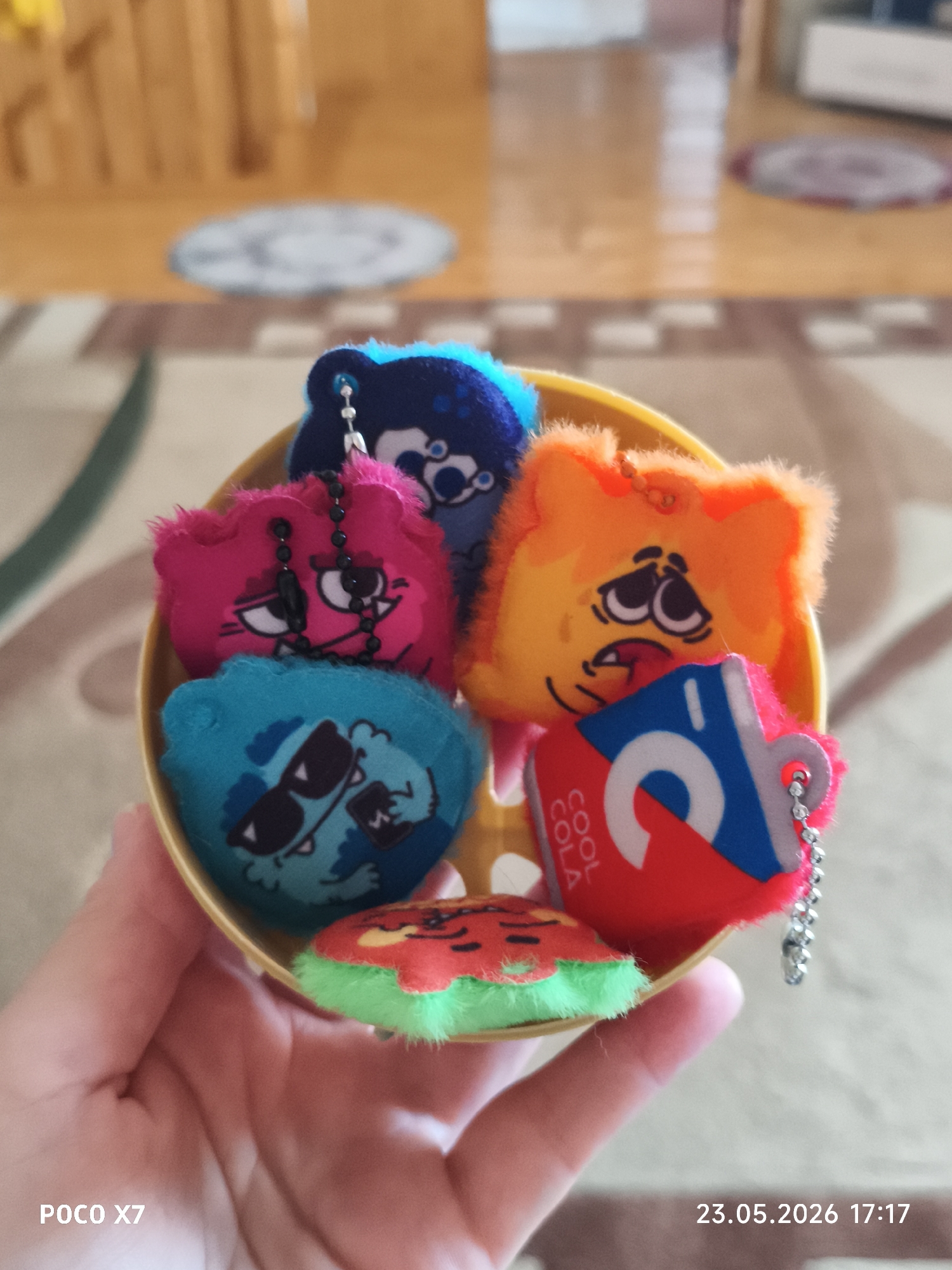 A close-up shot depicts a person holding a small, yellow plastic bowl containing six colorful plush keychains. The keychains include a blue fuzzy monster, a pink fuzzy monster, an orange fuzzy monster, a blue monster wearing sunglasses, a can of Cool Cola, and a slice of watermelon. The background is slightly blurred, showing a carpeted floor and furniture. The image includes a date and time stamp: "23.05.2026 17:17" and "POCO X7". Without additional information or context, the location cannot be definitively determined as Chaikovsky, Russia.