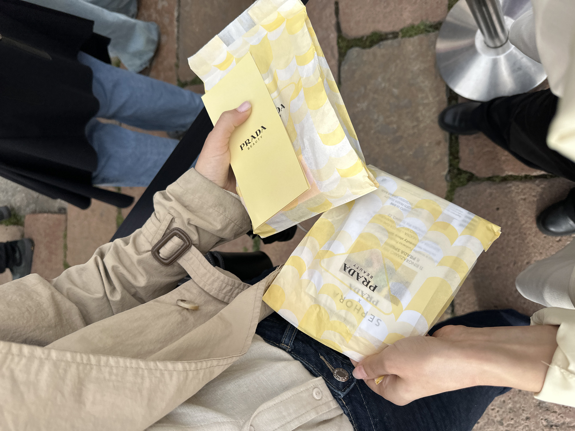 The scene depicts a person holding two yellow and white Prada Beauty x Sephora gift bags and a yellow Prada Beauty card in their hands. They are wearing a beige trench coat over a light-colored shirt and dark blue jeans. The location is suggested as Milan, Italy based on text on the bag that states, "TI RINGRAZIAMO PRADA SP," and "NUOVA-ZAGAMO, 22." The person is standing or sitting outdoors, with other people and a metal stanchion visible in the background. The ground appears to be paved with stone bricks.
