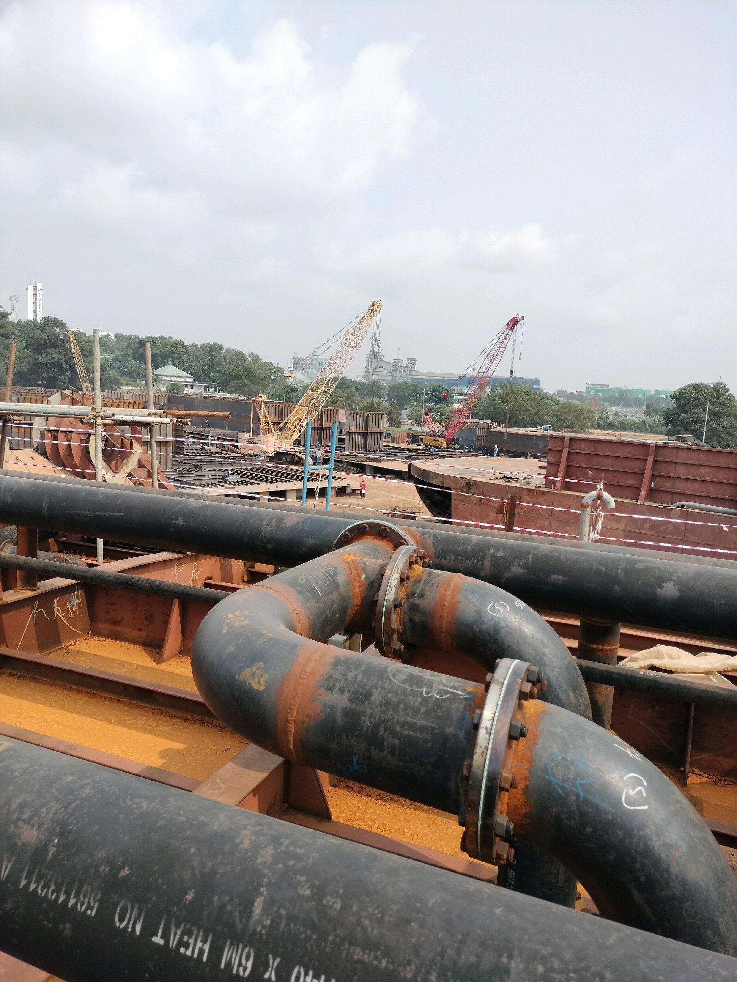 An aerial shot captures a sprawling shipyard in Batam, Indonesia, under a hazy sky. The foreground is dominated by large, dark, industrial pipes, some showing signs of rust and wear, connected with flanges and bolts. These pipes curve and snake across the scene, hinting at a complex network of infrastructure.
In the middle ground, the shipyard bustles with activity. Several large cranes, one yellow and one red, stand tall, their booms extended over sections of a massive ship or offshore structure under construction. Workers, appearing as small figures, can be seen scattered throughout the site, engaged in various tasks. Large, reddish metal plates and structural components are being assembled, forming the skeleton of a new vessel. Scaffolding and temporary supports are also visible, indicating ongoing construction.
The background reveals more industrial structures, including what appear to be factories and possibly a tall tower. Lush green trees form a dense treeline behind the shipyard, contrasting with the man-made environment. The overall impression is one of large-scale industrial activity and construction in a coastal city.