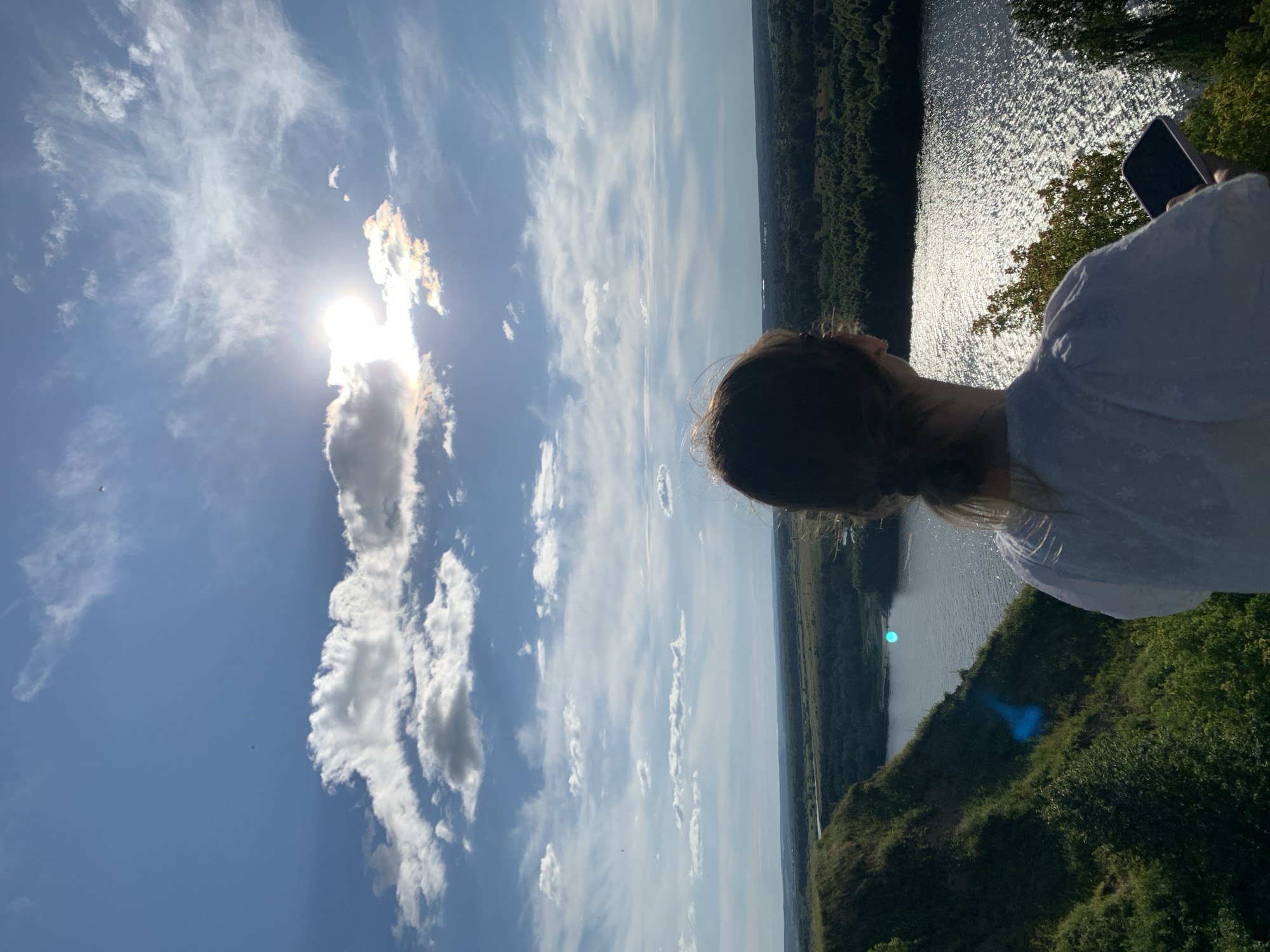 This image depicts a young woman from behind, looking out over a wide river and a lush green landscape. She is wearing a white top and her hair is light brown. In her right hand, she is holding a smartphone, suggesting she is either taking a picture or watching something on it.
The scene is bathed in bright sunlight, with the sun peeking through a scattered arrangement of white and wispy clouds, creating a somewhat hazy but beautiful sky. The sun's rays are reflecting brilliantly off the surface of the river, creating a shimmering effect. The river is bordered by dense, dark green trees that form a continuous line along the opposite bank and in the foreground. The immediate foreground shows a grassy, sloped area, also covered in greenery.
The time of day appears to be late afternoon or early evening, given the angle of the sun and the warm lighting. The weather is clear and sunny, with a gentle breeze suggested by the wispy clouds. The overall mood conveyed is one of peaceful contemplation and enjoyment of nature. The location, identified as Pavlovo, Russia, suggests a natural, possibly rural or park-like setting. There is no visible text in the image.