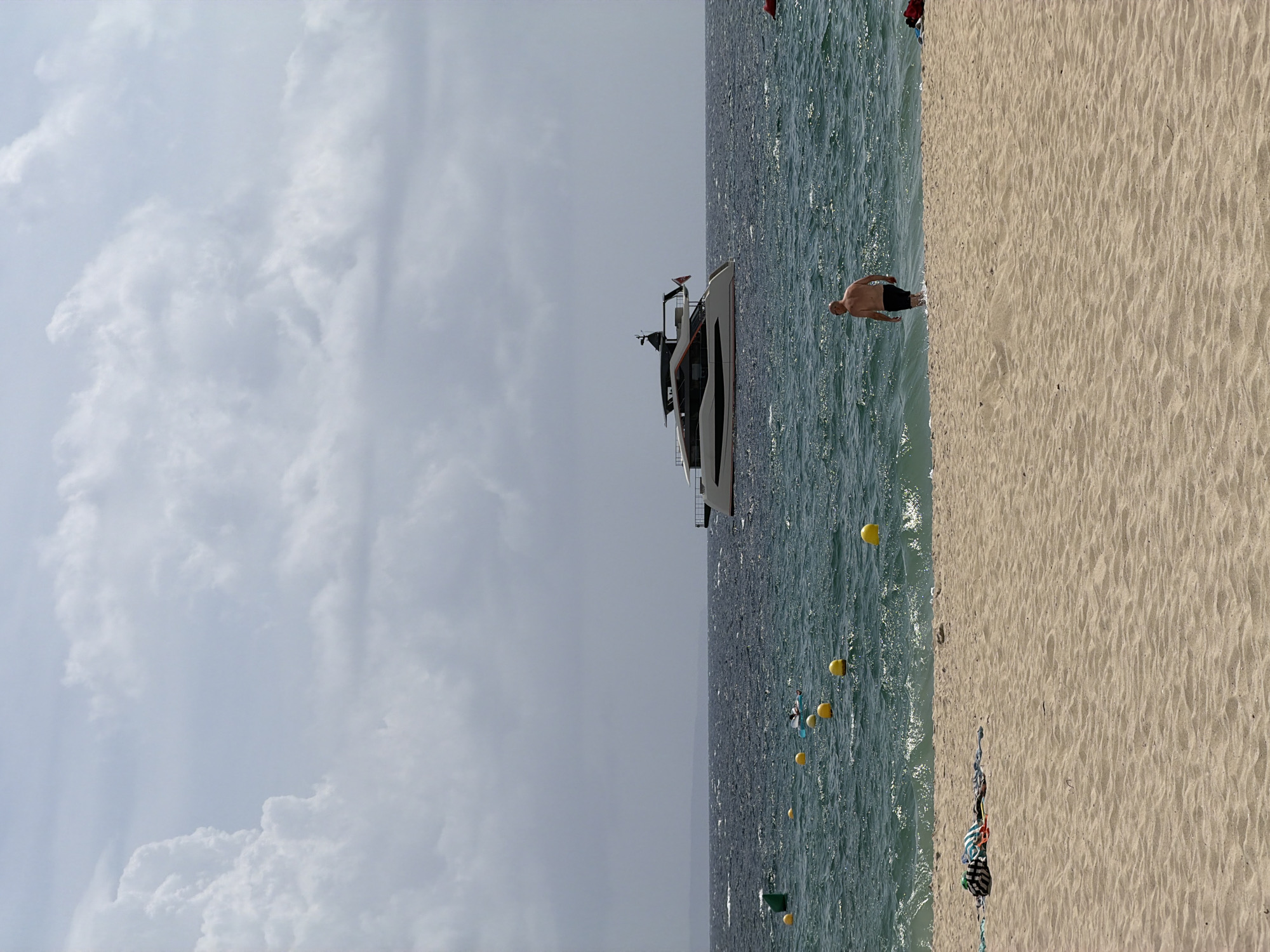 A man is walking into the ocean on a sandy beach in Palmanova, Spain. He is wearing black shorts. The water is choppy with small waves. Yellow buoys are floating in the sea. A large white yacht with black accents is docked in the distance. The sky is cloudy with a mix of white and gray clouds. On the beach, towels and a bag are laid out on the sand.