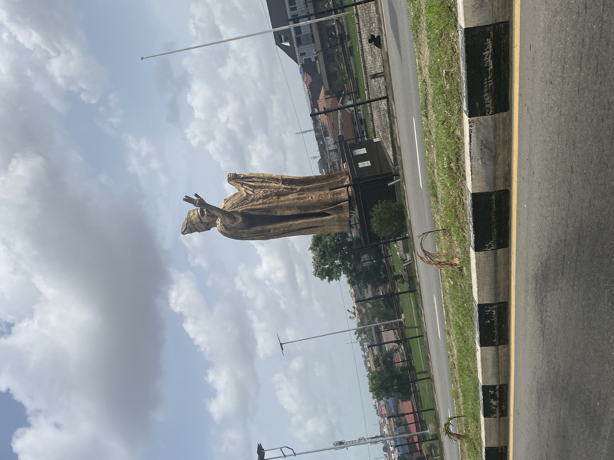 The image features a large, golden-hued statue of a human figure, likely a man, standing prominently outdoors. The statue is depicted with both arms raised and hands gesturing, possibly in a pose of blessing, welcome, or oratorical address. It appears to be wearing flowing robes or traditional attire.
To the right of the statue, a multi-lane asphalt road is visible, bordered by a black and white striped curb. A bright yellow line runs along the edge of the road, adjacent to the curb. Beyond the curb is a strip of green grass, followed by a black metal fence. In the background, several multi-story buildings with modern architecture are discernible, interspersed with numerous trees and other vegetation. Utility poles or lampposts are also visible in the distance and midground. The sky above is partially cloudy, with patches of blue showing through the white and gray clouds, indicating it is daytime. A slender flagpole is visible extending into the sky from the upper right. The scene suggests a public space, possibly a monument or a significant landmark, situated within an urban or suburban environment. There is no visible text in the image.