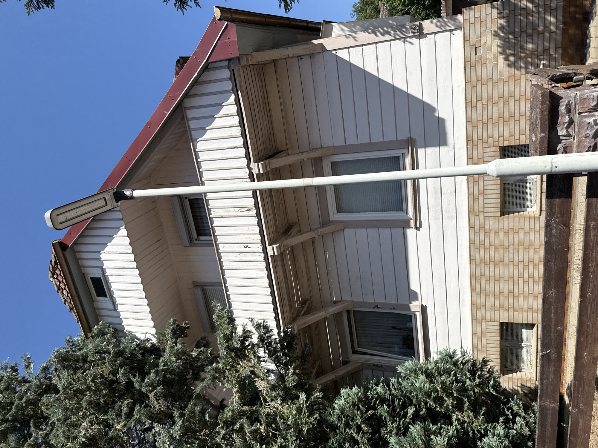 This is a medium-angle, eye-level, outdoor shot of the side of a two-story house with a prominent white wooden facade and a red metal roof. The house has a balcony on the upper level, supported by wooden beams. There are several windows visible, some with blinds. A tall, modern street lamp stands in front of the house, casting a shadow. To the left, a large, dark green evergreen tree partially obscures the house. The base of the house is constructed of light brown brick. A wooden fence is visible in the foreground. The sky is a clear, bright blue. The scene appears to be located in Bad Lauterberg im Harz, Germany.