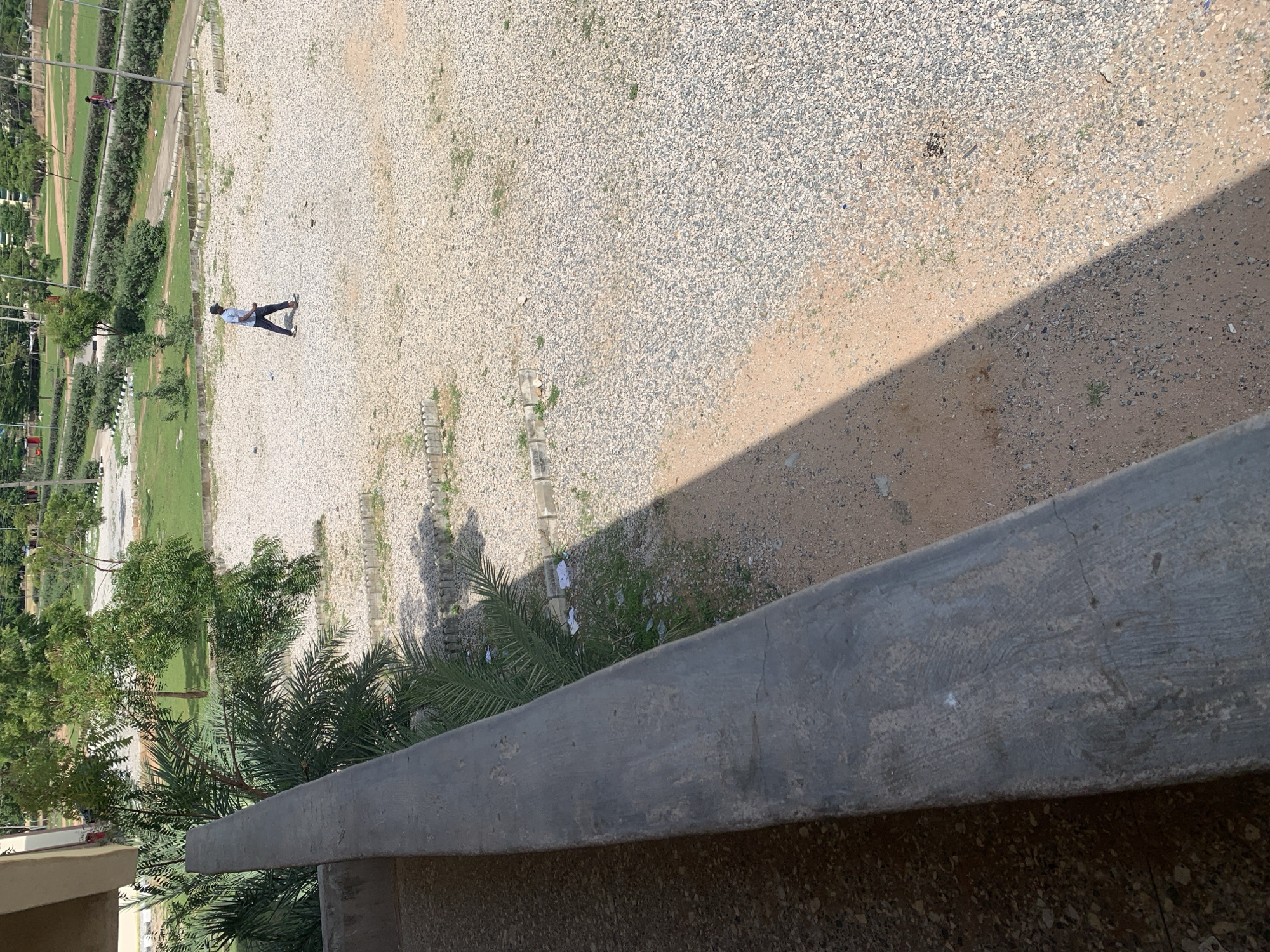 The image depicts an outdoor scene in the city of Jalingo, Nigeria, viewed from an elevated position. In the foreground, a rough, concrete railing runs along the left side of the frame, casting a sharp shadow across a sandy and gravelly area. To the left of the railing, a building structure is partially visible.
The midground shows a large expanse of light-colored gravel and sand, sparsely populated with small green plants. In the center of this area, a person is walking. They are wearing a light-colored t-shirt, dark trousers, and dark shoes. They are walking from left to right in the frame, their posture suggesting a casual stride. Further in the background, there is a neatly manicured lawn with scattered trees and bushes. Beyond the green spaces, there are visible structures and what appear to be utility poles with wires. Another person is seen walking on a paved path in the far background, to the right of the frame. The overall environment suggests a park or an open area within an urban setting, characterized by a mix of natural and man-made elements. The lighting indicates it is daytime, with the sun casting strong shadows.