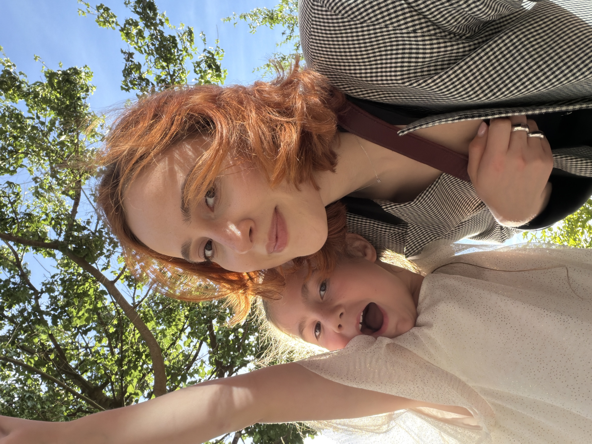 The image shows a close-up selfie of two females, a young girl and an adult woman, taken outdoors under a tree with green leaves. The adult woman has red hair and is wearing a black and white checkered jacket and a thin silver necklace. She is holding her left hand near her chest, revealing two silver rings on her left ring finger. The young girl, positioned to the left of the woman, has blonde hair and is wearing a sparkly white dress. Her mouth is open as if she is speaking or laughing, and her left arm is raised. The background consists of a bright blue sky and the branches of a tree. The lighting suggests it is daytime. There are no specific landmarks indicating the location is in Strasbourg, France.