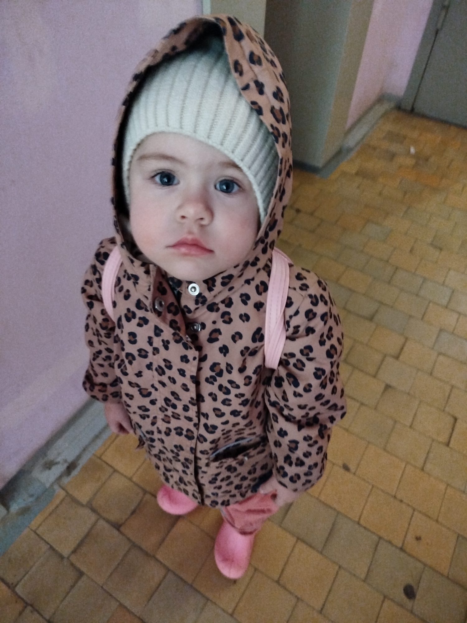 A young child is standing on a tile floor. The floor appears to be in the interior of a building. The child is wearing a beige beanie underneath a hooded animal print coat and pink boots. The child is looking at the camera. The location is Chaikovsky, Russia.