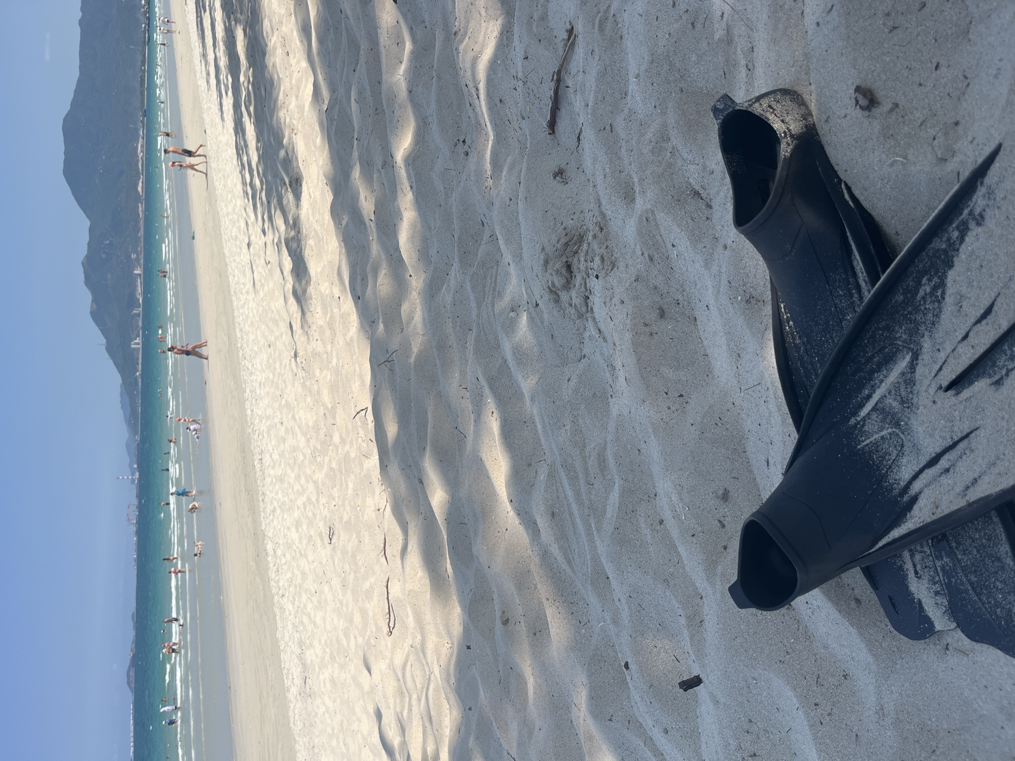 The image shows a pair of black swim fins lying on a sandy beach. In the background, there is a coastline with people in the water and on the sand. A mountain range is visible in the distance. The sky is clear and blue. The location is Unknown, Unknown.