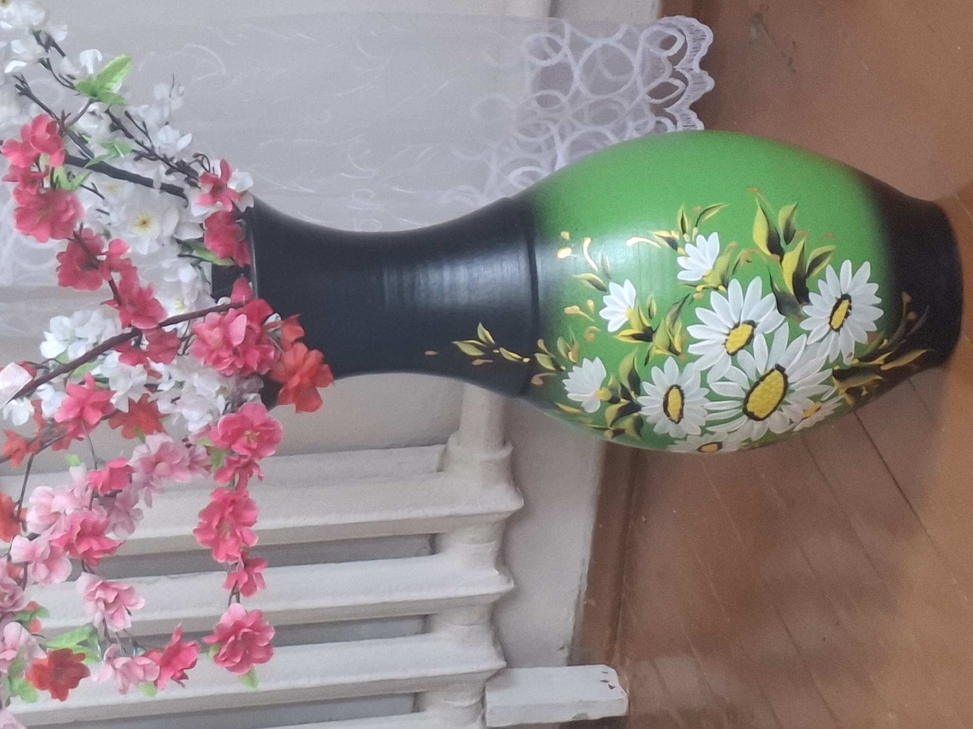 A wide shot captures a black and green ceramic vase, adorned with painted white daisies and green leaves, placed on a wooden floor. The vase is filled with artificial flowers in shades of pink and white, resembling cherry blossoms. In the background, a white radiator with vertical slats is visible, suggesting an indoor setting. A white sheer curtain with a circular pattern hangs on the right side of the image. The floor displays a warm, polished wood grain. There are no people or animals present.