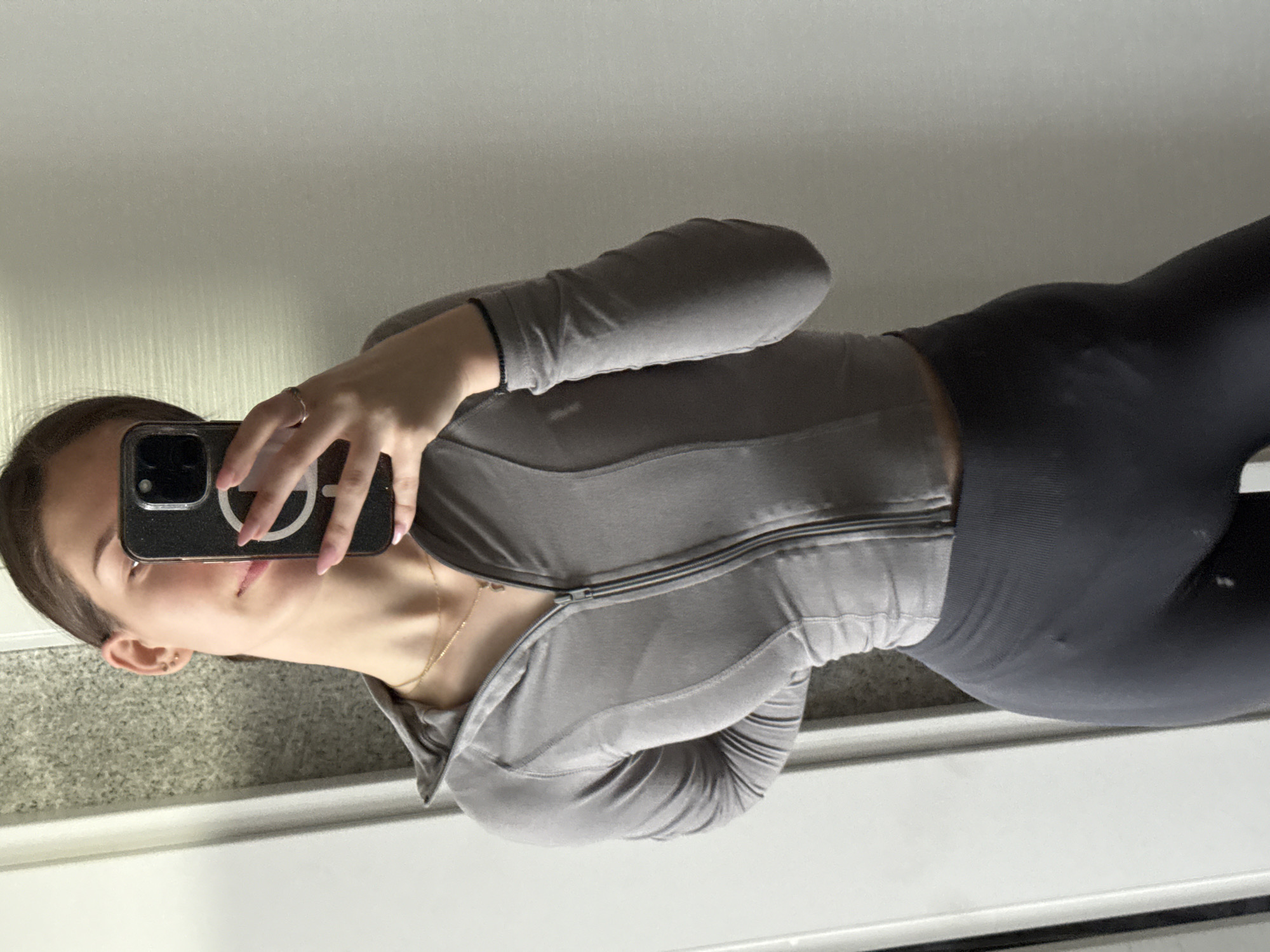 A woman takes a selfie in a mirror. She is wearing a gray athletic zip-up jacket and dark gray leggings. She is holding a black iPhone with a white circular emblem on the back. A gold necklace is visible around her neck. The reflection shows a tiled wall and white trim, suggesting an interior space, possibly a bathroom or dressing area. There is no information about the location in Sofia, Bulgaria.