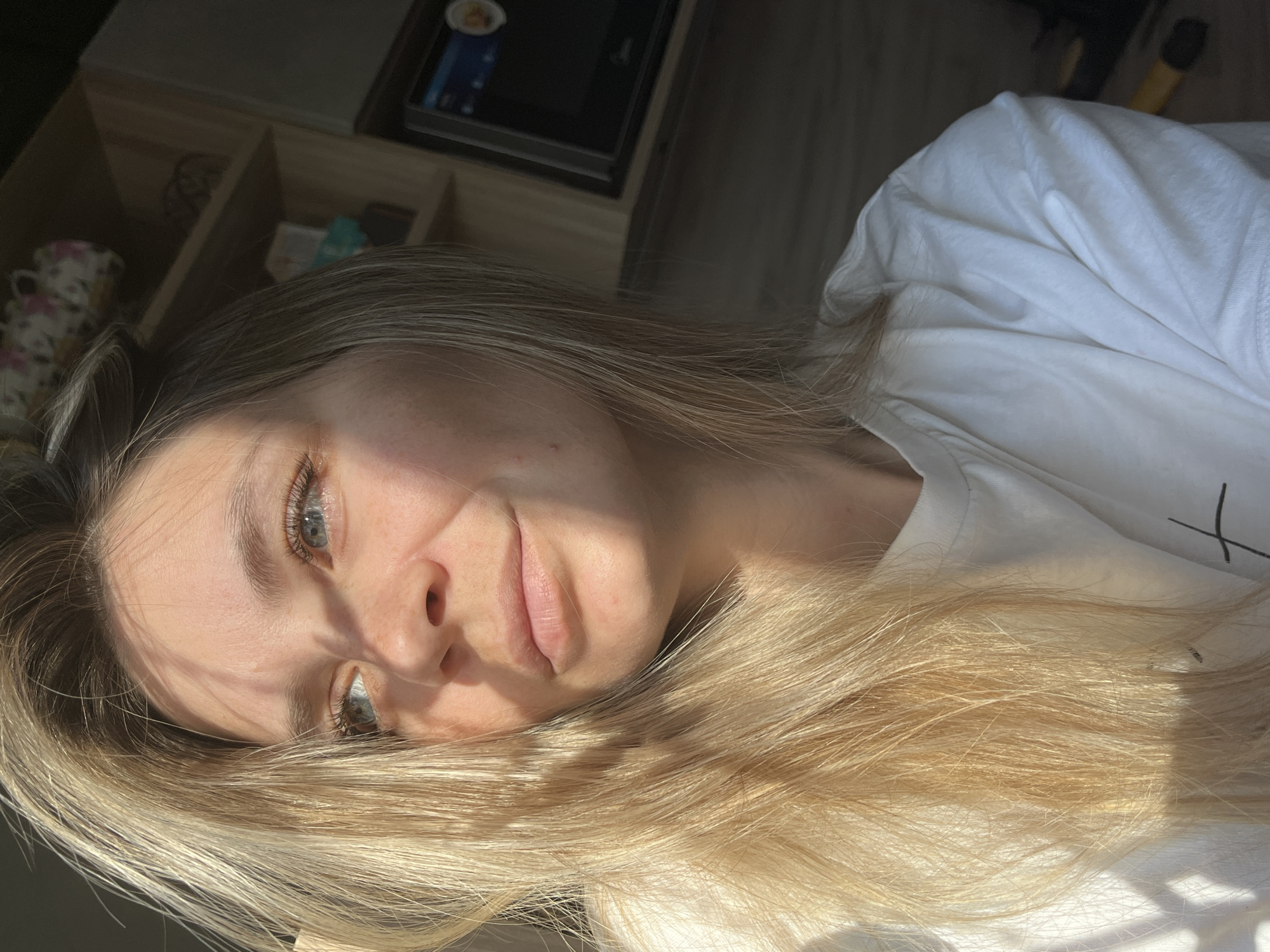 A young woman with long, light blonde or ash-blonde hair and striking light-colored eyes (blue-green) is captured in a close-up, upward-angled portrait. She is lying down, possibly on a bed or floor, in what appears to be an indoor setting in Chaikovsky, Russia. Her head is tilted, and her hair is spread out around her, catching the strong, directional sunlight that creates prominent shadows across her face and body. This suggests the time of day is either morning or late afternoon.
The woman is wearing a simple white t-shirt, which has some indistinguishable dark markings on it, possibly part of a graphic or print. Her expression is calm and somewhat pensive, as she looks directly towards the viewer. The natural light highlights her skin and the texture of her hair.
In the blurred background, shadowed by the strong light, elements of a domestic environment are visible. These include a wooden shelving unit or drawer with multiple compartments, a white mug with a floral pattern, and what looks like a dark electronic device, possibly a mobile phone or tablet. No decipherable text is visible anywhere in the image. The overall impression is one of quiet repose bathed in natural light.
