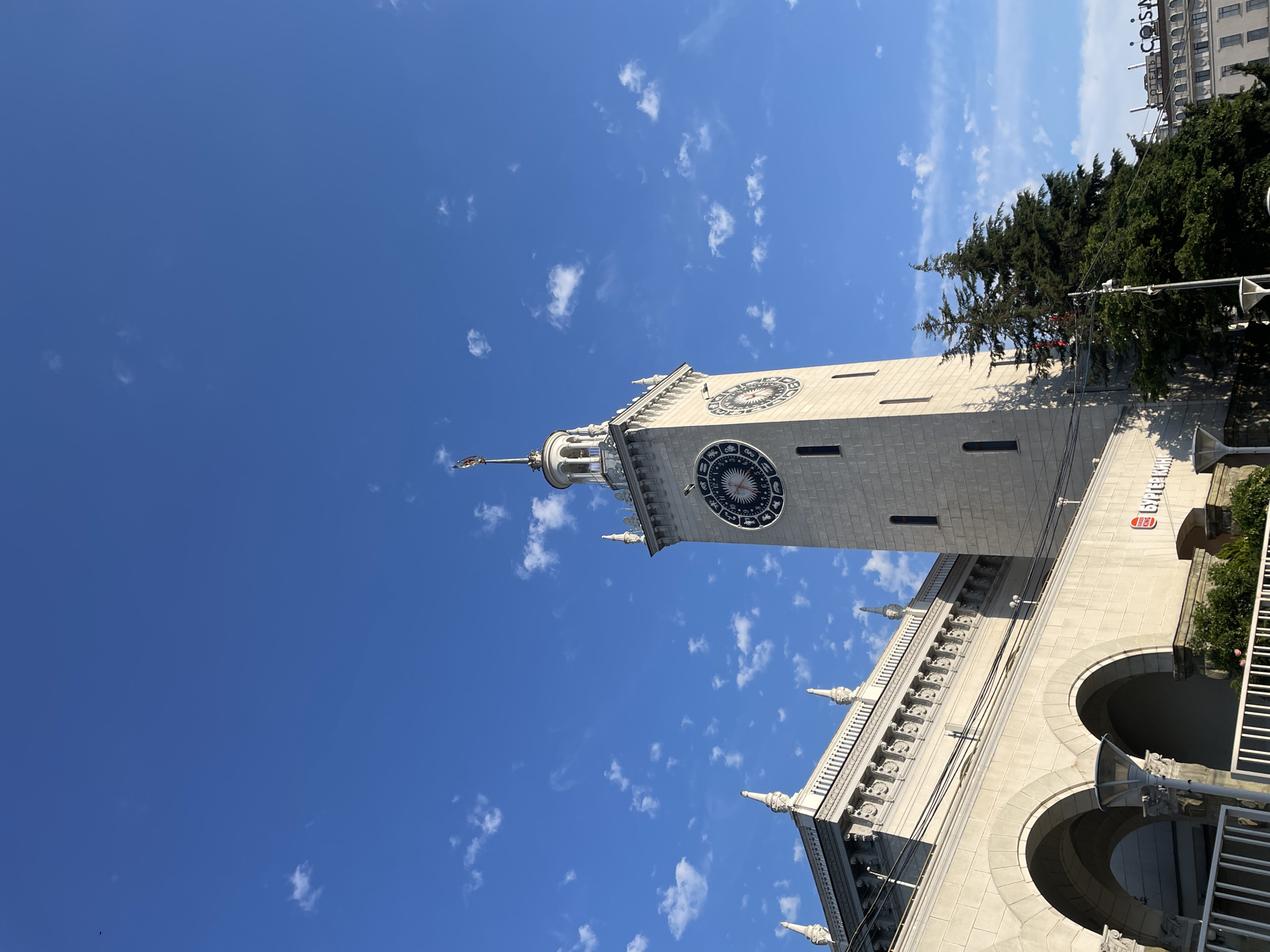 The image captures a low-angle view of the Sochi Sea Station, a prominent landmark in Sochi, Russia. The building, constructed from light-colored stone, features an ornate facade with arched entrances and decorative elements along the upper levels. A tall clock tower, also made of stone, rises above the main structure, displaying a circular clock face with astrological symbols. The sky is a vibrant blue, dotted with small, wispy white clouds, suggesting a clear and sunny day. A tall evergreen tree stands to the right of the clock tower, its dark green foliage contrasting with the bright sky and pale stone building. In the background, the upper floors of another building are visible, with the word "COCOA" in large white letters on a dark background. The overall scene conveys a sense of grandeur and architectural beauty, characteristic of public buildings in a seaside resort. The time of day appears to be midday or early afternoon, given the bright sunlight casting shadows. No people are visible in the frame.