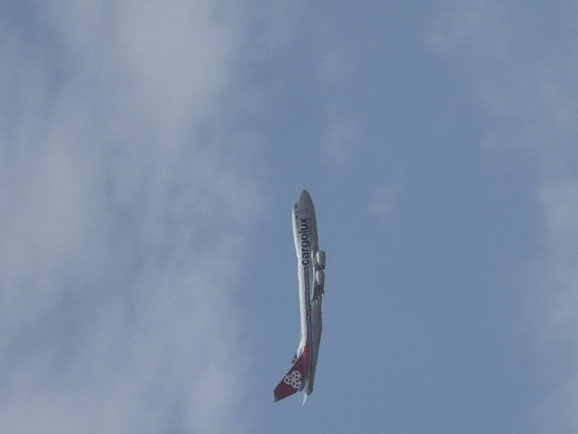 An airplane flies in the sky with clouds. The airplane is white with red and blue stripes along its side. The tail of the plane has a red logo with white accents. The word "cargolux" is written in blue on the side of the plane. The sky is a light blue with white clouds scattered throughout. The scene is likely in the vicinity of Mondercange, Luxembourg, as that is where Cargolux, Luxembourg's national airline, is headquartered and operates from.