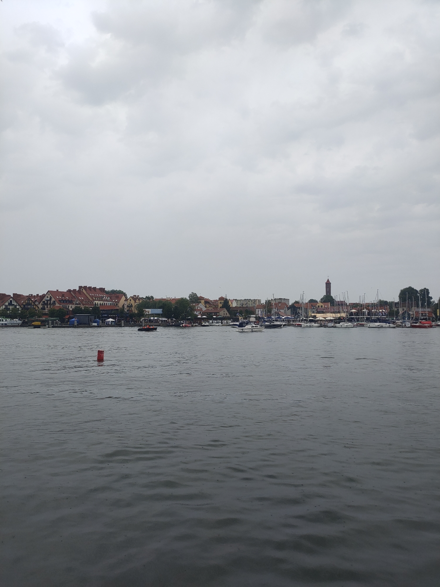 The image depicts a cloudy day over the town of Mikołajki, Poland, with a view of the harbor and Masurian Lake. In the foreground, the dark water of the lake is rippled, with a single red buoy visible. The middle ground showcases a waterfront lined with buildings, trees, and a variety of boats docked in the harbor. These include sailboats with tall masts, motorboats, and a yellow barge. A small orange and white boat with several people on board is moving on the water. In the background, the town's skyline features buildings with red-tiled roofs and a prominent clock tower. The sky is overcast with a uniform layer of gray clouds, creating a subdued atmosphere.