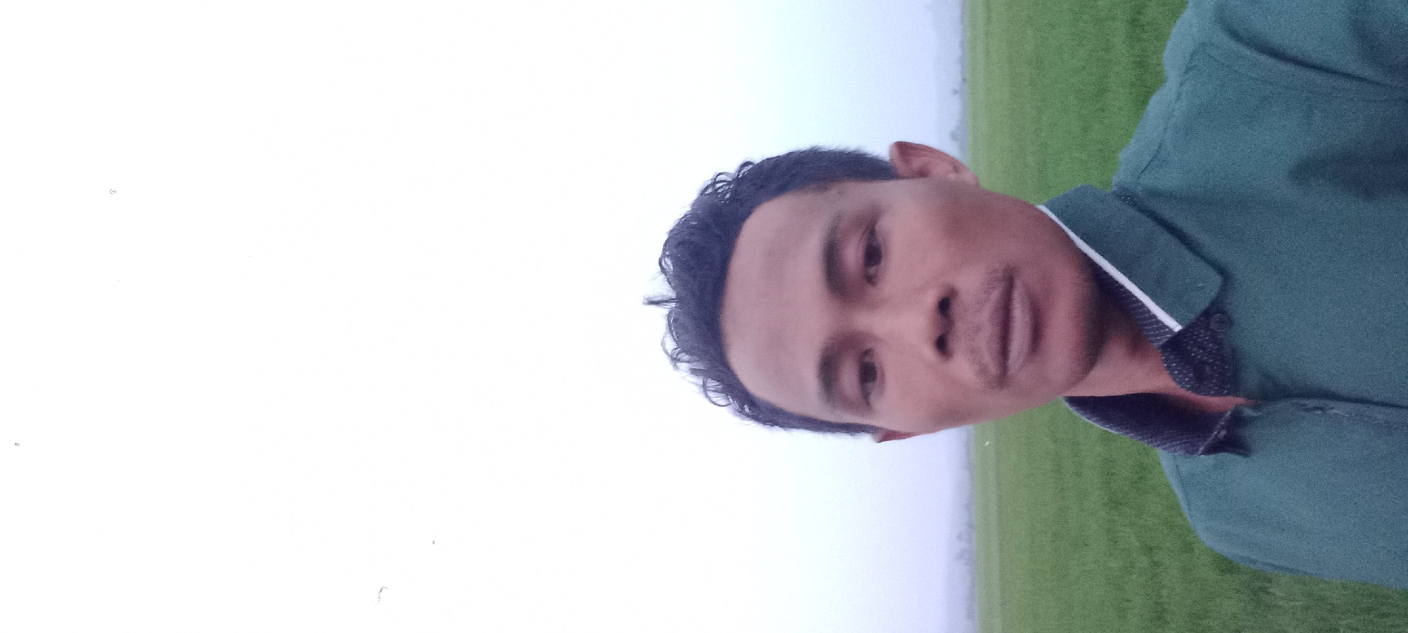 The image shows a close-up, selfie-style shot of a man in his late 20s or early 30s. He has dark, curly hair and is wearing a dark green collared shirt with a white and black striped detail on the collar. He is standing outdoors with a blurry green field behind him, suggesting a rural or agricultural setting. The sky is overcast and light, creating a diffused light source. The man's expression is neutral. The location is identified as Sidamulya, Indonesia.