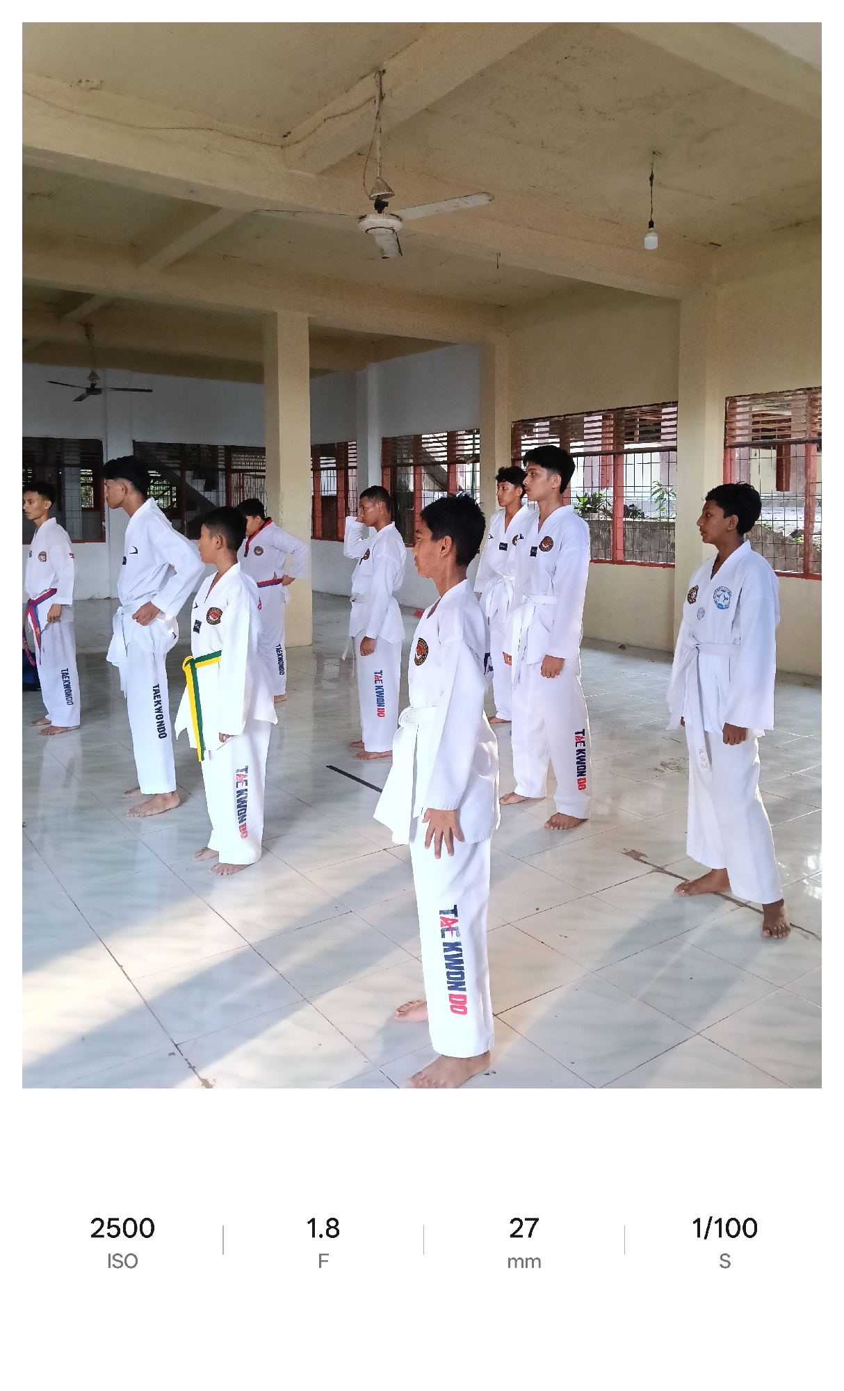 Certainly, let's analyze the image.
**Scene:** The photograph appears to be taken indoors, likely in a gymnasium or large hall with tiled flooring. The location is in Lhokseumawe, Indonesia.
**People:** There's a group of individuals, mostly young boys, wearing white martial arts uniforms (taekwondo). Their uniforms are adorned with the words "TAEKWONDO" and "TAE KWON DO". Some are wearing colored belts (yellow and others). All of the people appear to be barefoot.
**Objects:** Visible within the space are pillars, windows with bars, ceiling fans, and an overhead light fixture.
**Actions:** The group appears to be standing in formation, likely in the middle of a training session or a practice exercise.
**Environment:** The hall seems to be well-lit, with sunlight coming through the windows. The walls appear to be painted in a light beige color.