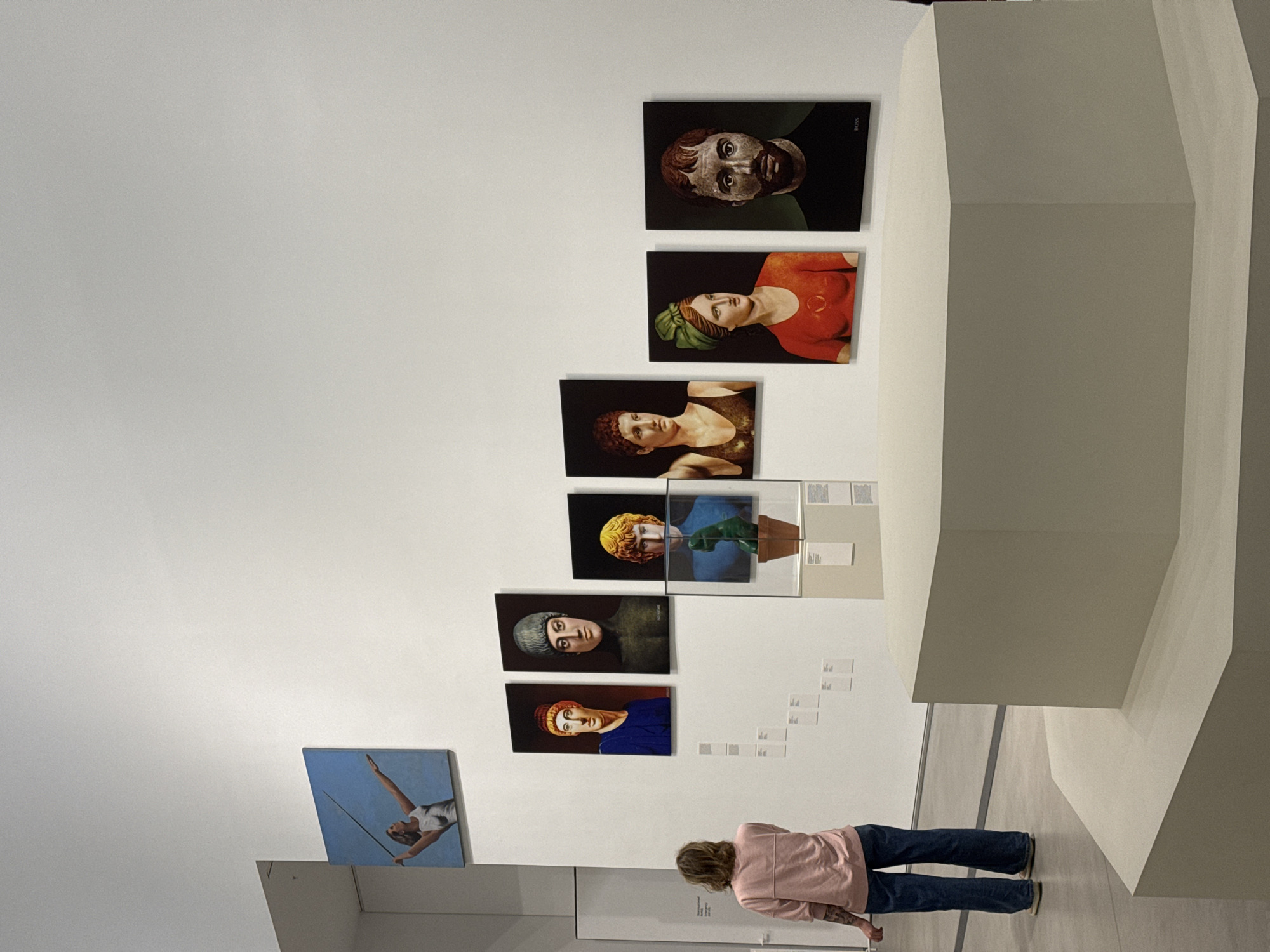 An image taken in an art gallery or museum shows a person observing a series of artworks displayed on a bright white wall. The original image is rotated 90 degrees clockwise.
Seven portraits are arranged vertically on the wall, depicting individuals from the chest up, with a classical or stylized appearance against dark backgrounds. These portraits feature strong colors, and the word "BOSS" is visible vertically printed on the right side of at least two of the canvases. Below these, another artwork on the far left shows a person in a white outfit against a light blue background, appearing to be an athlete or performer holding a long, thin object.
In the foreground, a person with medium-length hair, wearing a pink long-sleeved top and blue jeans, is seen from behind, standing on a light-colored floor and facing the art displays. To the right of the artworks, a large, angular, light-grey architectural structure or installation forms a prominent feature of the room. Small white labels are visible on the wall below some of the artworks, and transparent protective cases are positioned in front of a few pieces, suggesting an exhibition setting. The overall aesthetic is clean and modern, characteristic of a contemporary art space. The scene is indoors, likely lit by artificial light. The activity is art viewing. The location context provided is Moscow, Russia.