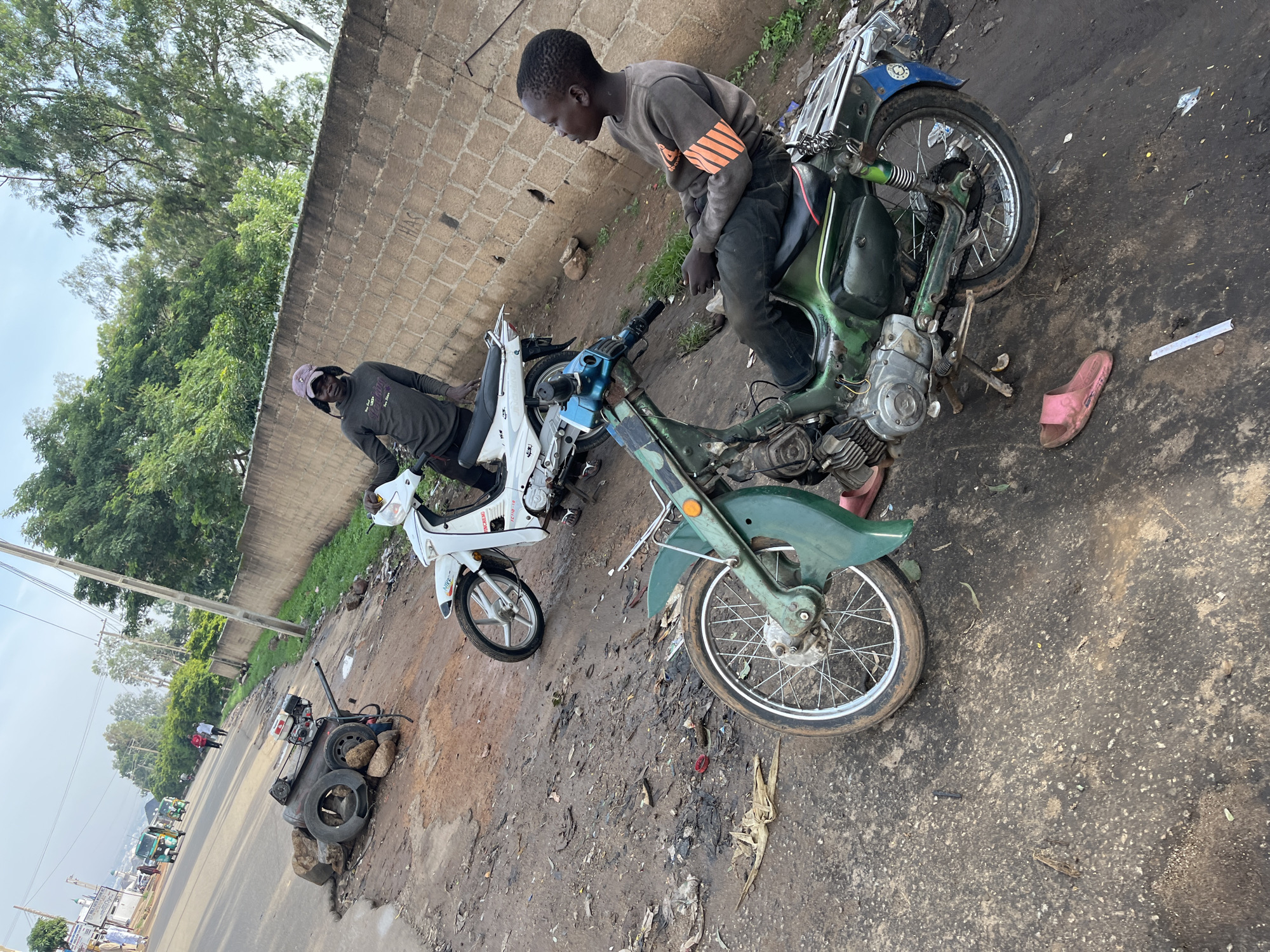 This image captures a roadside scene in Jos, Nigeria, with two young boys near parked motorcycles. In the foreground, a boy sits on a green motorcycle. He wears a dark sweatshirt with orange stripes on the sleeves and dark pants. A single pink sandal lies on the ground near the motorcycle. Behind him, a white motorcycle is parked. A second boy, wearing a dark long-sleeved shirt and a purple baseball cap, stands next to the white motorcycle. To the left of the motorcycles, a disassembled piece of machinery, possibly a generator, is placed on the ground next to two tires and rocks. The environment appears to be a dusty, unpaved roadside with scattered debris and a brick wall in the background. Trees are visible beyond the wall, and the sky is overcast. In the distance, on the paved road, there are moving vehicles, including a three-wheeled taxi.