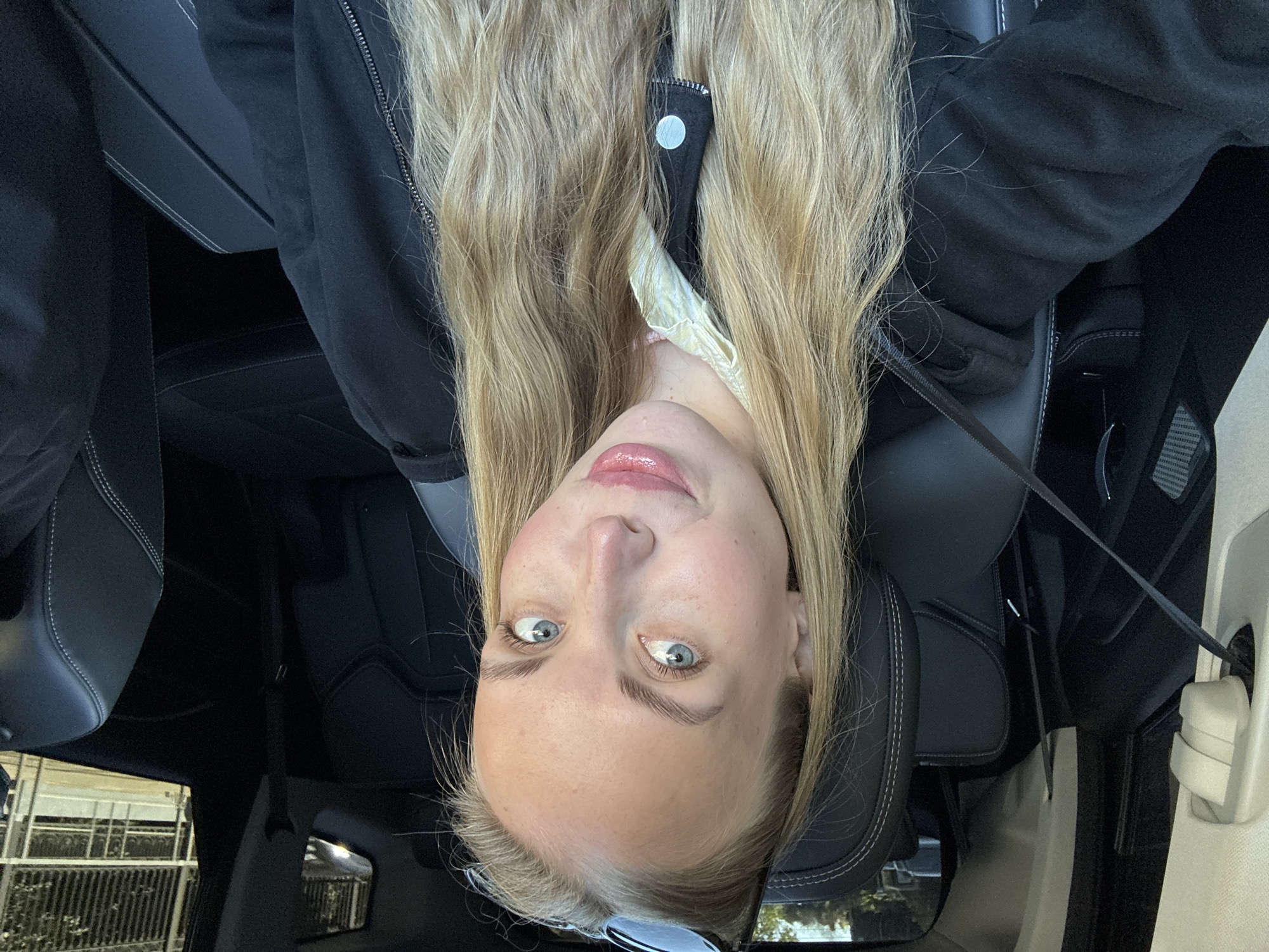 This media file presents an inverted full-face portrait of a young woman situated inside a car, likely in Sochi, Russia. The woman has long, straight blonde hair that falls around her face and shoulders. Her light blue eyes are wide and directly focused on the viewer, and her lips are slightly parted, adorned with a glossy, light pink lip product. She possesses fair skin and prominent eyebrows.
She is wearing a dark, possibly black, jacket or hoodie, with a zipper detail visible near her neck. A lighter-colored fabric, which could be from an undergarment or scarf, is visible beneath her jacket. A seatbelt strap diagonally crosses her upper body, suggesting she is seated in a moving vehicle.
The car's interior features black leather seats with distinct white stitching. Elements of the dark dashboard or door panels are also visible. Through the car windows, a blurred outdoor environment can be seen, including green foliage on one side and a structure with horizontal bars, resembling a fence or building, on the other. The lighting indicates it is daytime. No visible text is present in the image.