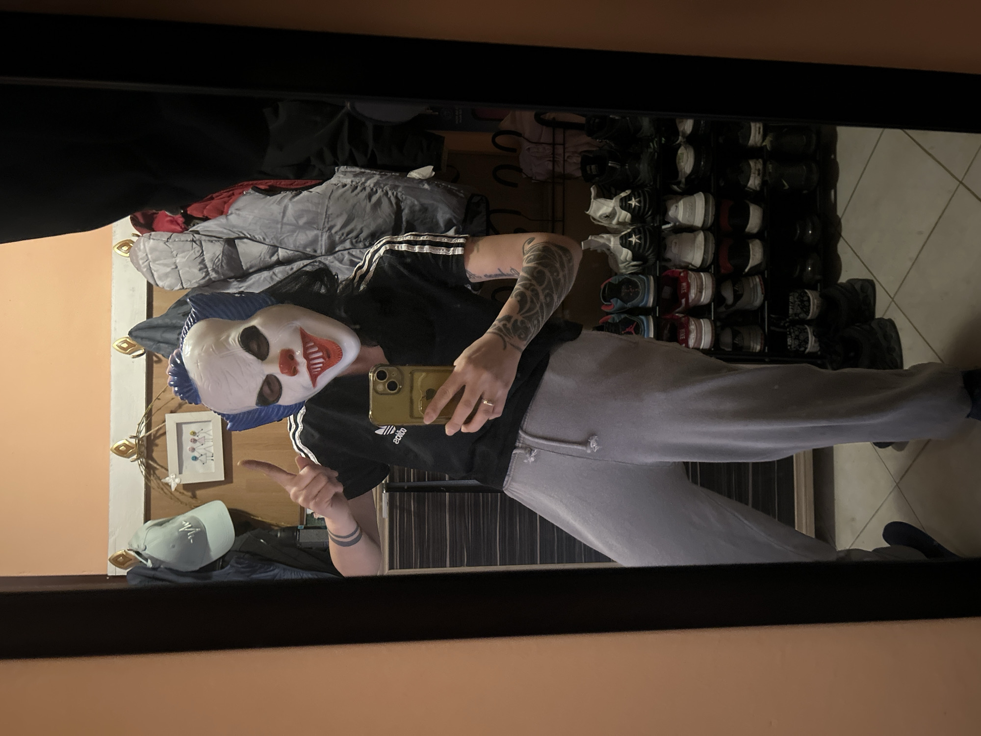 A person wearing a clown mask, an Adidas t-shirt, and grey sweatpants is standing in front of a mirror, holding a phone. The person is making a gesture with their left hand. Behind the person is a closet filled with clothes and shoes. The location is Unknown, Unknown.