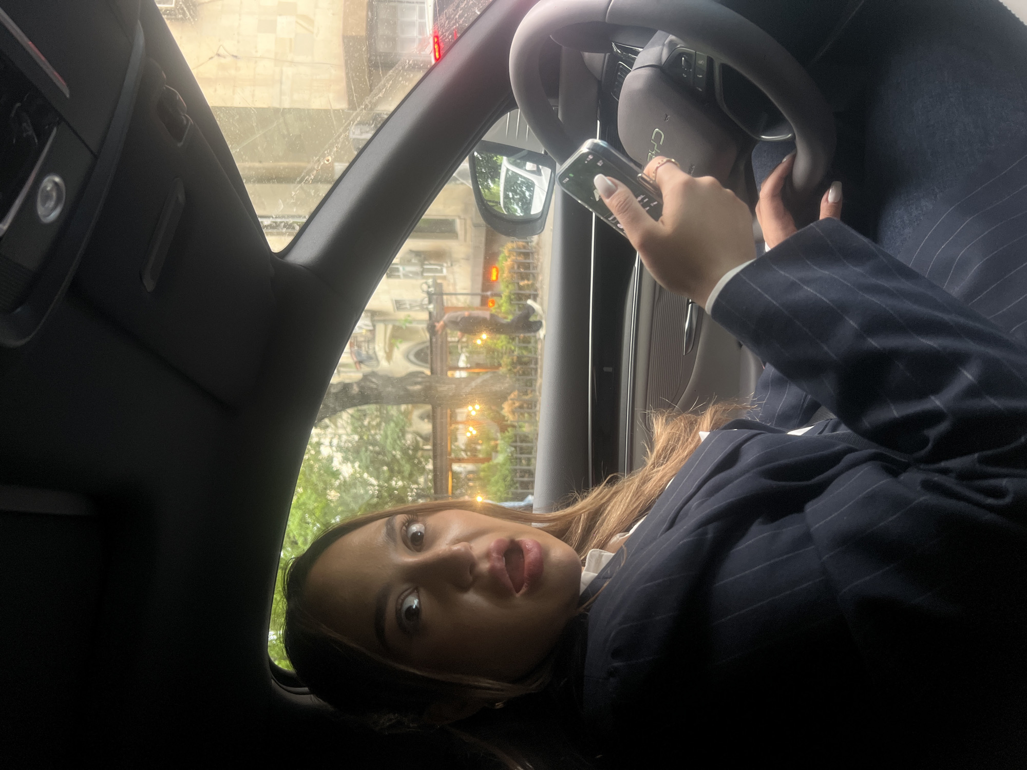 A woman in a pinstripe suit is seated in the driver's seat of a car, holding a smartphone in her right hand near the steering wheel. The view from the car window shows buildings with a light color scheme, indicating an urban environment. The photo was taken in Yerevan, Armenia.