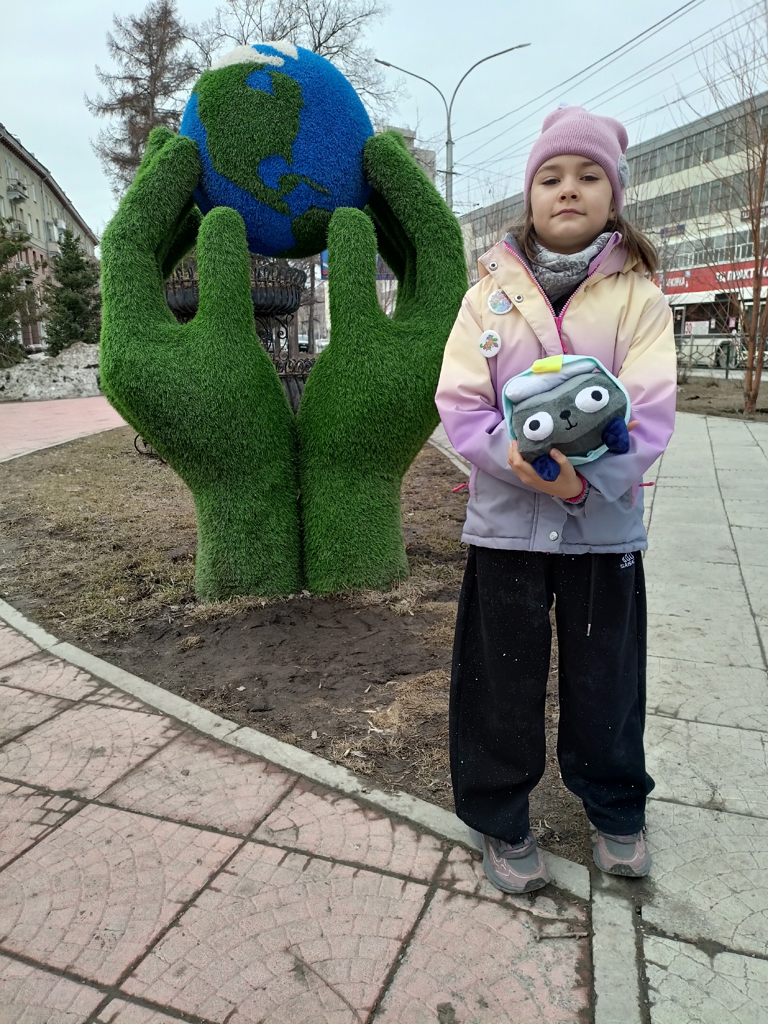 A young girl stands on a sidewalk in what appears to be a city park or public space in Novosibirsk, Russia. She is holding a plush toy resembling a character, possibly from a video game. Behind her is a large decorative sculpture of two green hands holding a globe. The surrounding environment includes buildings, bare trees, and patches of snow, suggesting it is a cold season. The sidewalk is made of patterned concrete tiles.