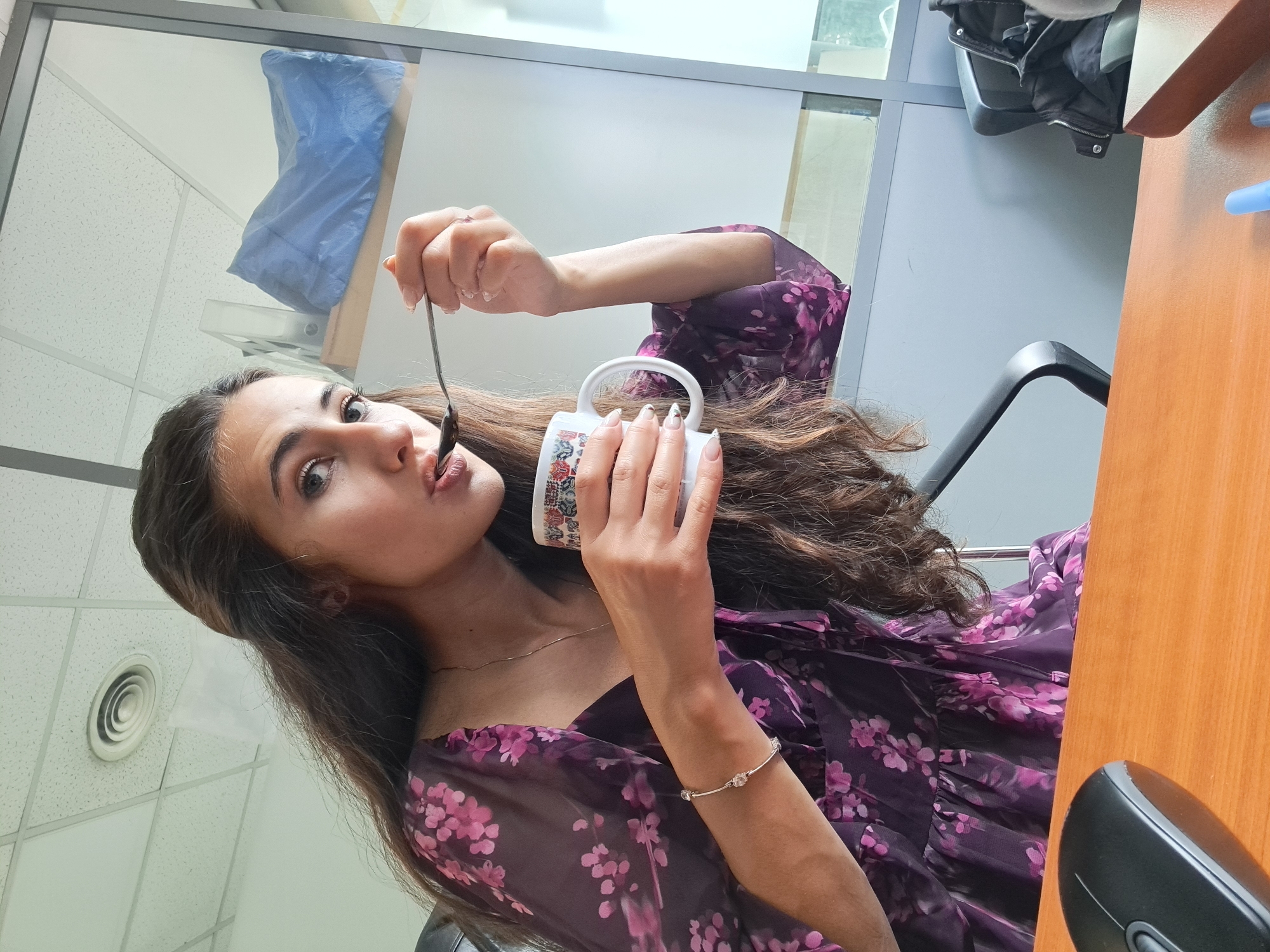 A young woman is captured in a close-up, upward-angled shot, appearing to eat or taste something from a white mug with a spoon. Her head is tilted back, and her eyes are looking towards the camera with an engaged expression. She has long, dark, wavy hair cascading around her. She is wearing a dark purple dress with a vibrant pink floral pattern. A silver-colored bracelet with sparkling elements adorns her left wrist, and her nails are painted with a light-colored polish featuring a subtle design.
In her right hand, she holds a metal spoon containing a dark substance, which is positioned at her lips. Her left hand holds the white mug, which has a colorful, intricate pattern on its side.
The setting appears to be an indoor office or commercial space. Visible in the background is a white ceiling with a grid pattern and a circular air vent. Behind her, glass partitions with metal frames separate spaces, and a blue, possibly plastic-covered object or bag is seen behind one of the glass panels. To her right, a wooden desk surface is visible, along with part of a black computer mouse. A black office chair can also be seen behind her. The overall lighting suggests an interior environment, possibly during the daytime. No text is visible in the image.