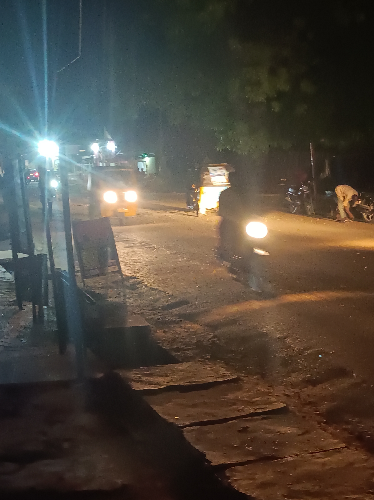 The image depicts a dimly lit street scene in Narayi, Nigeria, at night. A yellow tuk-tuk is visible in the middle distance, its headlights illuminating the road. A motorcycle with a single headlight approaches closer to the camera. Several parked motorcycles are seen in the background to the right, where a person bends over one of the vehicles. Streetlights cast a stark glow, and shadows are long and pronounced. The road surface appears rough and uneven. A sign and some metal structures stand on the left side of the road.