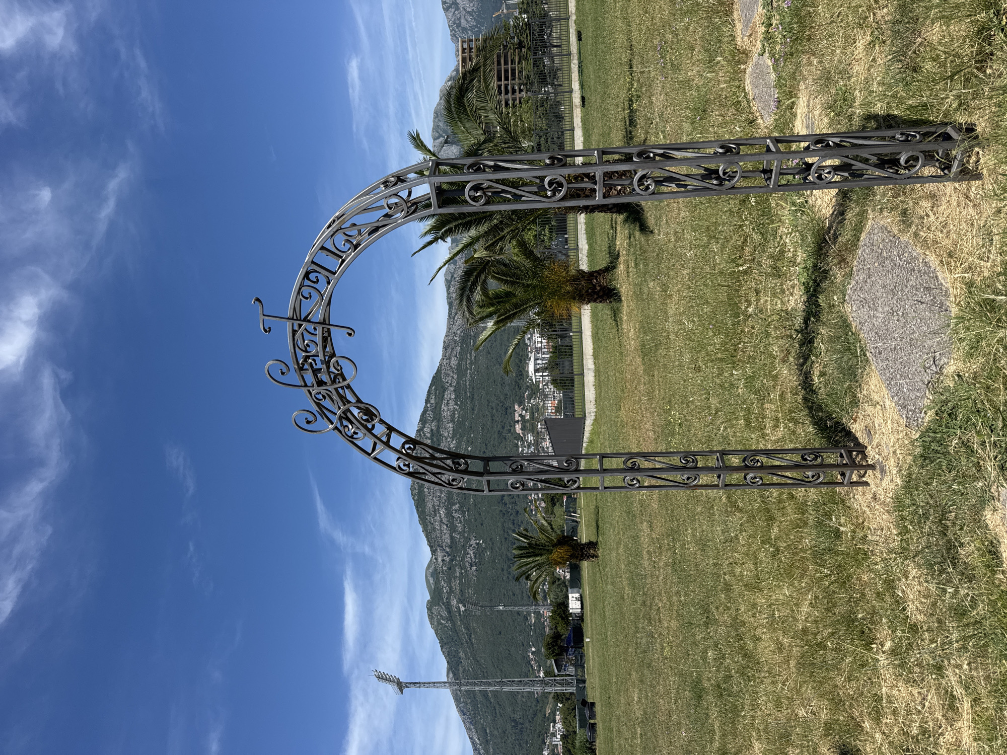 The image depicts a decorative metal archway in a grassy area with a view of a mountain and part of the city of Bar, Montenegro, visible in the background. The archway features ornate, wrought-iron-style patterns. Palm trees are interspersed within the view, adding a tropical feel to the Montenegrin landscape. The sky is a clear blue with light, wispy clouds. There's no immediate sign of people in the frame, focusing on the composition of the landscape and the decorative structure.