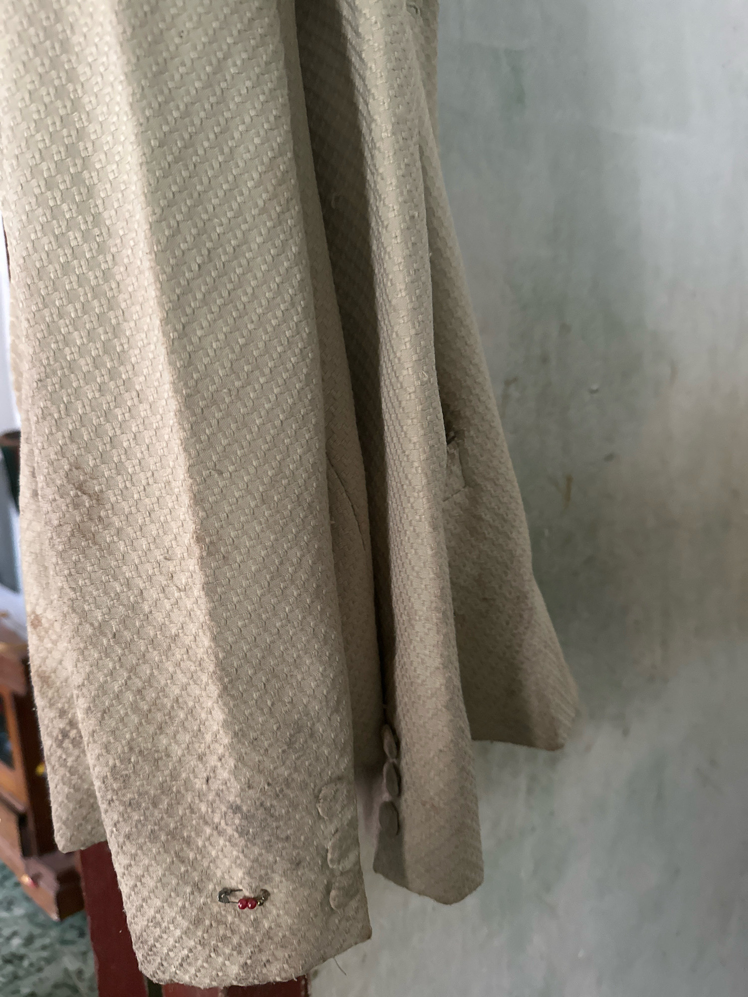 A close-up, eye-level shot focuses on the textured fabric of a tan jacket or coat hanging on a wall in what appears to be a residential setting in Margamulya, Indonesia. The fabric has a distinct woven pattern. On the left sleeve cuff, a small safety pin with two red beads attached is visible. The jacket is draped, with folds and creases creating shadows and highlighting the texture. The background is a textured, light-colored wall with some green smudges, possibly water stains or paint. The overall lighting is soft, suggesting an indoor environment with diffused natural light.