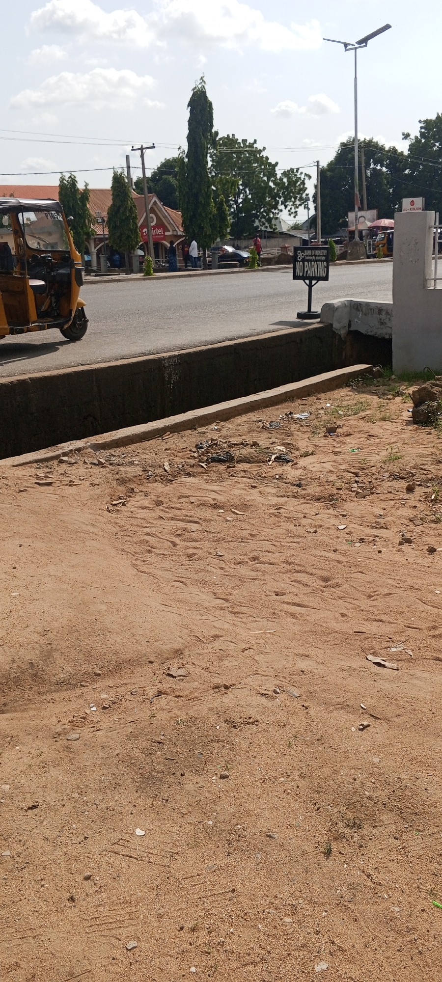 The image depicts a street scene in Jalingo, Nigeria. In the foreground, a sandy area with scattered debris and tire tracks is visible, bordering a concrete drainage ditch. Beyond the ditch, a paved road runs horizontally across the frame. On the far side of the road, several buildings with red roofs are present, including one with an "Airtel" sign. A yellow auto-rickshaw is parked on the left side of the road. A black and white sign displaying "BANK OF INDUSTRY NO PARKING" stands on the right side of the road, near a lamppost. Several individuals are seen walking or standing near the buildings. The sky is partly cloudy.