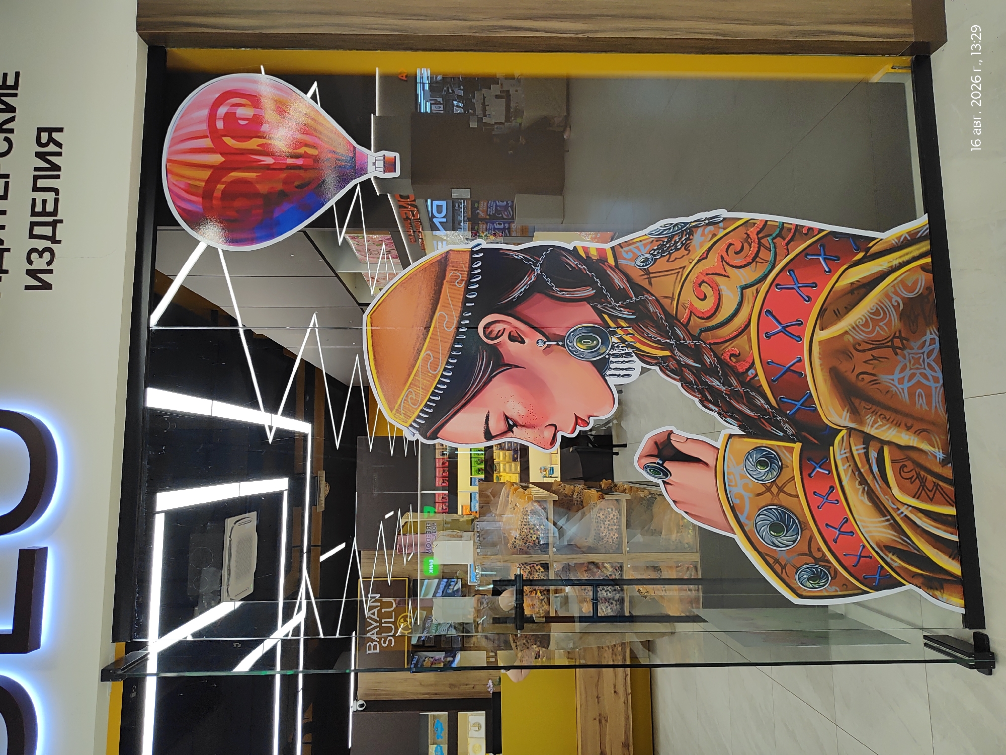 The image captures a storefront in Astana, Kazakhstan, featuring a large, stylized illustration of a young woman wearing traditional Kazakh attire and a hot air balloon. The artwork is affixed to the exterior of a glass entrance, suggesting it's a decorative element for a business.
The woman in the illustration is depicted in profile, her gaze directed downwards. She wears an orange and brown patterned garment adorned with blue cross-stitching and circular motifs. Her head is covered with a traditional cap, and she sports a large, ornate earring. A thick braid of dark hair is visible. She wears a ring on her finger.
Above and to the right of the woman, a colorful hot air balloon is illustrated. Its design features swirling patterns in shades of red, orange, yellow, and blue, with a visible heart shape incorporated into the design.
Through the glass storefront, the interior of a shop is visible. Shelves stocked with various products, possibly food items, can be seen. A sign with Cyrillic text, "СУЛУ" (SULU) is partially visible on the left side of the storefront. Another sign with Cyrillic text above the entrance reads "КОНДИТЕРСКИЕ ИЗДЕЛИЯ" (CONFECTIONERY PRODUCTS).
The foreground shows the tiled floor and the lower framing of the glass entrance. The overall impression is that of a vibrant, culturally inspired advertisement for a confectionery or retail business. The timestamp "16 авг. 2026 г., 13:29" is visible in the bottom right corner.