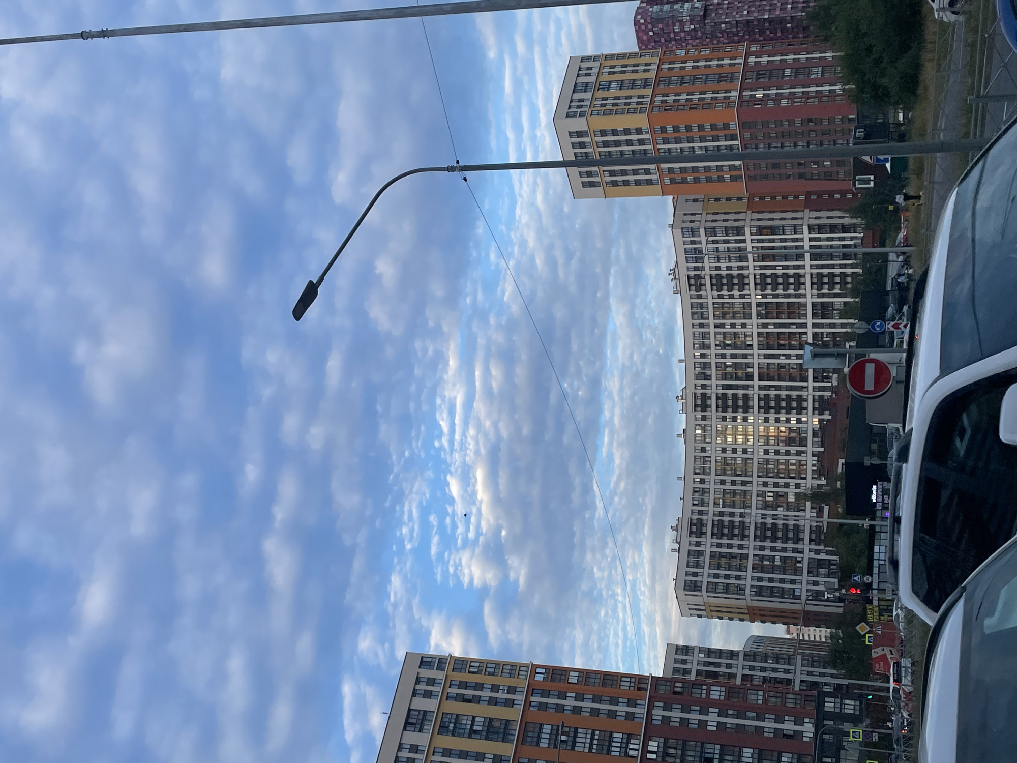 This image captures a street view in the Vyborgsky District of Saint Petersburg, Russia, under a cloudy sky. Dominating the scene are several tall, modern residential buildings with colorful facades. In the foreground, the tops of several cars are visible, suggesting a street-level perspective. A lamppost curves over the road, and power lines crisscross the sky. Behind the cars and trees, a large, curved residential building stands prominently, its windows reflecting the light. To its left and right are other tall apartment buildings, one predominantly orange and red, and another with a reddish-purple hue. A red circular sign with a white horizontal bar, indicating "no entry," is visible on the right side of the road. Several smaller signs and traffic lights are also present, indicating a busy urban environment. A few pedestrians can be seen walking on the sidewalk in the middle ground. The overall impression is of a contemporary urban landscape in a residential area.