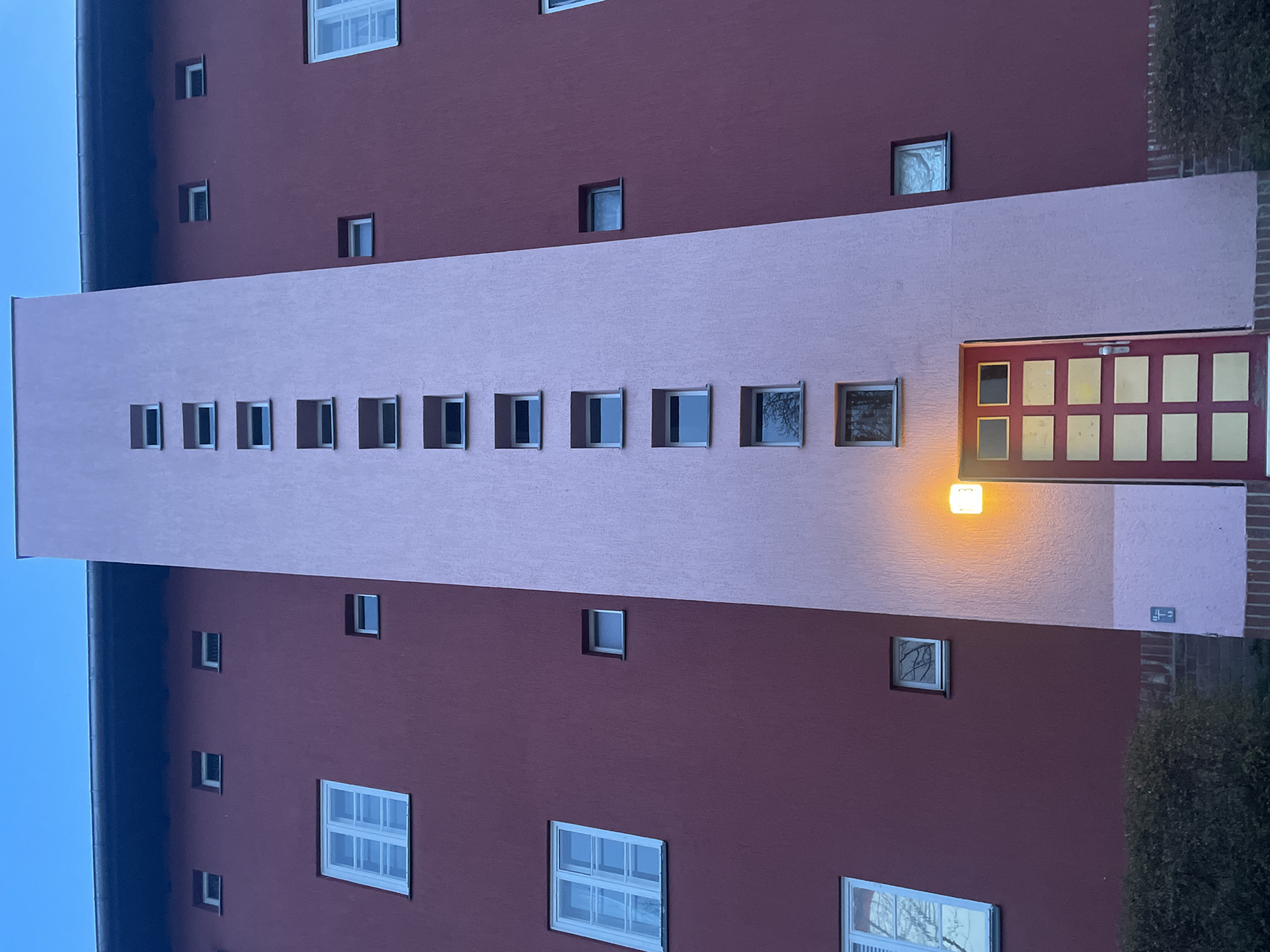 The image depicts an apartment building in Berlin, Germany. The building has a distinctive architectural style, featuring a reddish-brown facade with rectangular windows of varying sizes. A prominent, light pink vertical section runs up the center of the building. It has a series of small, square windows along its length. A door with a window in it is on the right, at the bottom of the vertical pink element. A streetlamp is visible above the door. The sky is visible at the top and left of the frame, with a twilight or dusk appearance. A small bush is on the right side of the image.