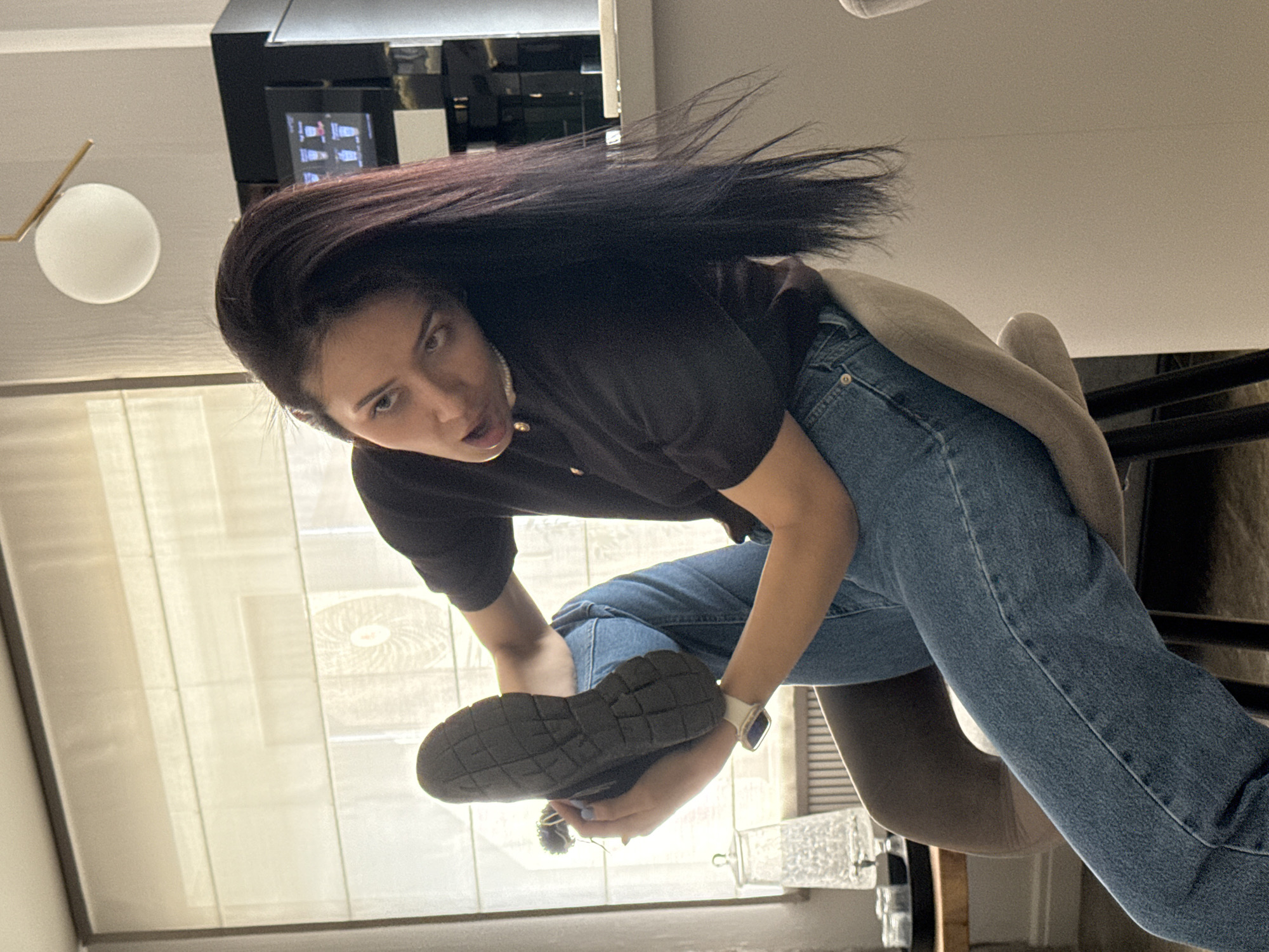 A woman is standing in an indoor setting. She is wearing a dark-colored t-shirt, blue jeans, and a watch. Her dark hair is partially blowing upward. In her hands, she holds a dark-colored shoe with a textured sole. Behind her are blinds and a window. To her left is a light fixture with a round, white shade. A black structure with a screen is visible behind her head. There is no visual evidence that the image was taken in Votkinsk, Russia.