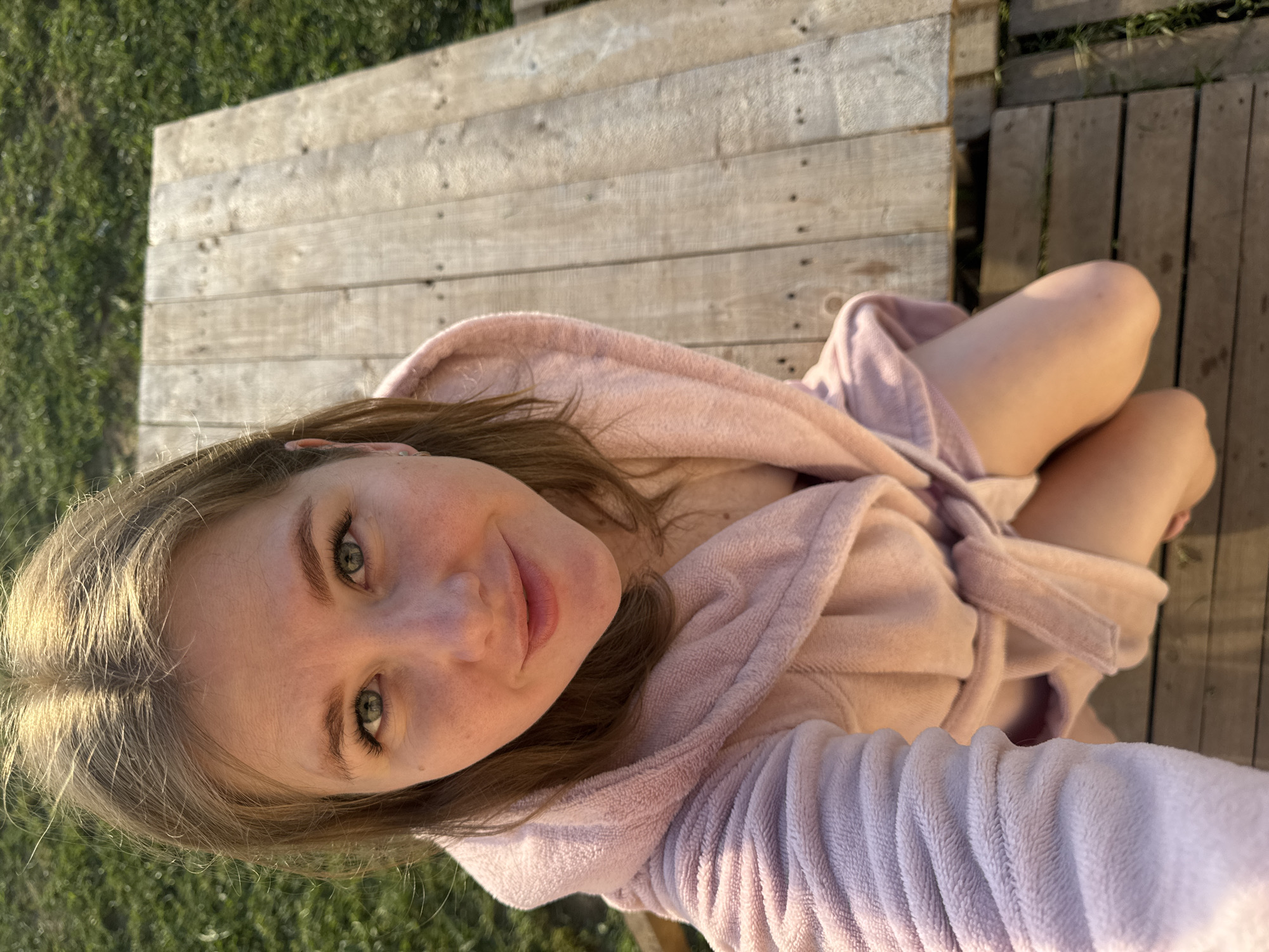 A young woman wearing a light pink robe is sitting on a wooden platform, with a grassy area visible on the edges of the platform. She is looking directly at the camera, smiling slightly. The photograph appears to have been taken from a high angle, looking down at her. The wooden platform is constructed of light-colored planks. The scene is outside, and likely in Ферма, Russia.
