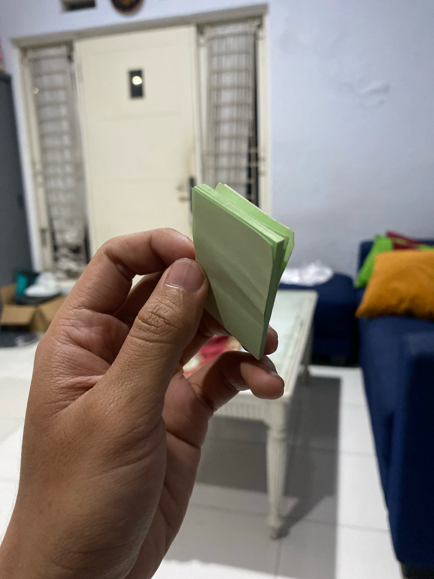 In Klurak, Indonesia, a person's hand is holding a small stack of light green paper, possibly sticky notes, angled towards the right. The background shows a room with a cream-colored door featuring a small dark rectangular window with a bright light inside. To the left of the door are vertical window grills. In the mid-ground, a white table with ornate legs is visible. Further to the right, a portion of a dark blue sofa with orange and green cushions is in the background. The floor appears to be tiled. The overall lighting suggests an indoor, daytime setting.