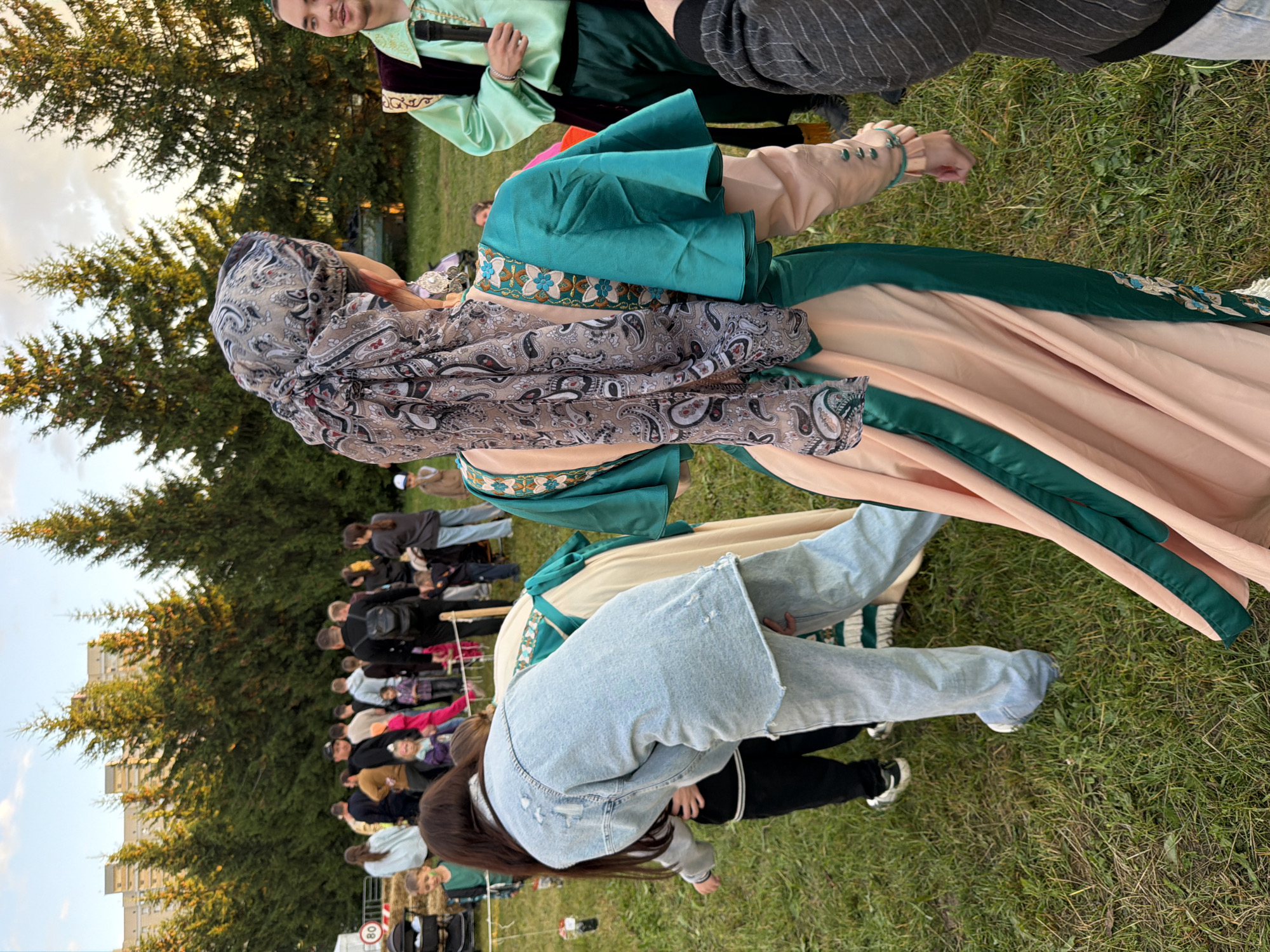 An eye-level, medium shot shows a group of people gathered outdoors in what appears to be a park or festival, with tall evergreen trees and modern, multi-story buildings in the background. The lighting suggests it is late afternoon, with warm, golden sunlight filtering through the trees. In the foreground, a person wearing a pale blue denim jacket and matching jeans is bending over, interacting with a child in black pants and a white-collared shirt. To their right, a woman in a traditional peach-colored dress with teal trim and a patterned headscarf is visible from the back, her arms outstretched as if moving. Another person in a bright green tunic and dark pants, holding a microphone, is partially visible on the right side of the frame. The ground is covered in green grass with some dry patches. A white rope barrier with a sign showing a red circle with a diagonal line and the number 80 is visible to the left, along with a white tent-like structure and hay bales. The overall atmosphere is casual and social, possibly at an outdoor event or gathering.