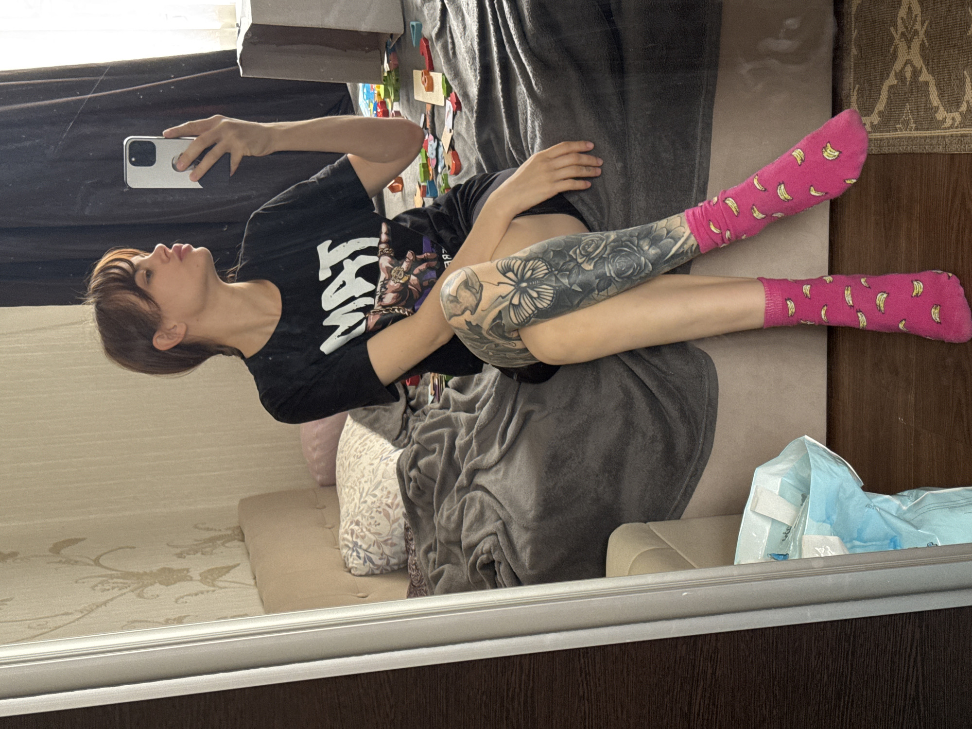 A woman with brown hair is taking a mirror selfie. She is lying on a bed covered with a grey blanket and patterned pillows. She wears a black t-shirt and pink socks with bananas. A visible tattoo adorns her leg. There is a pack of medical supplies next to the bed. The location may be a private residence in Rostov-on-Don, Russia, but this is only potentially inferred.