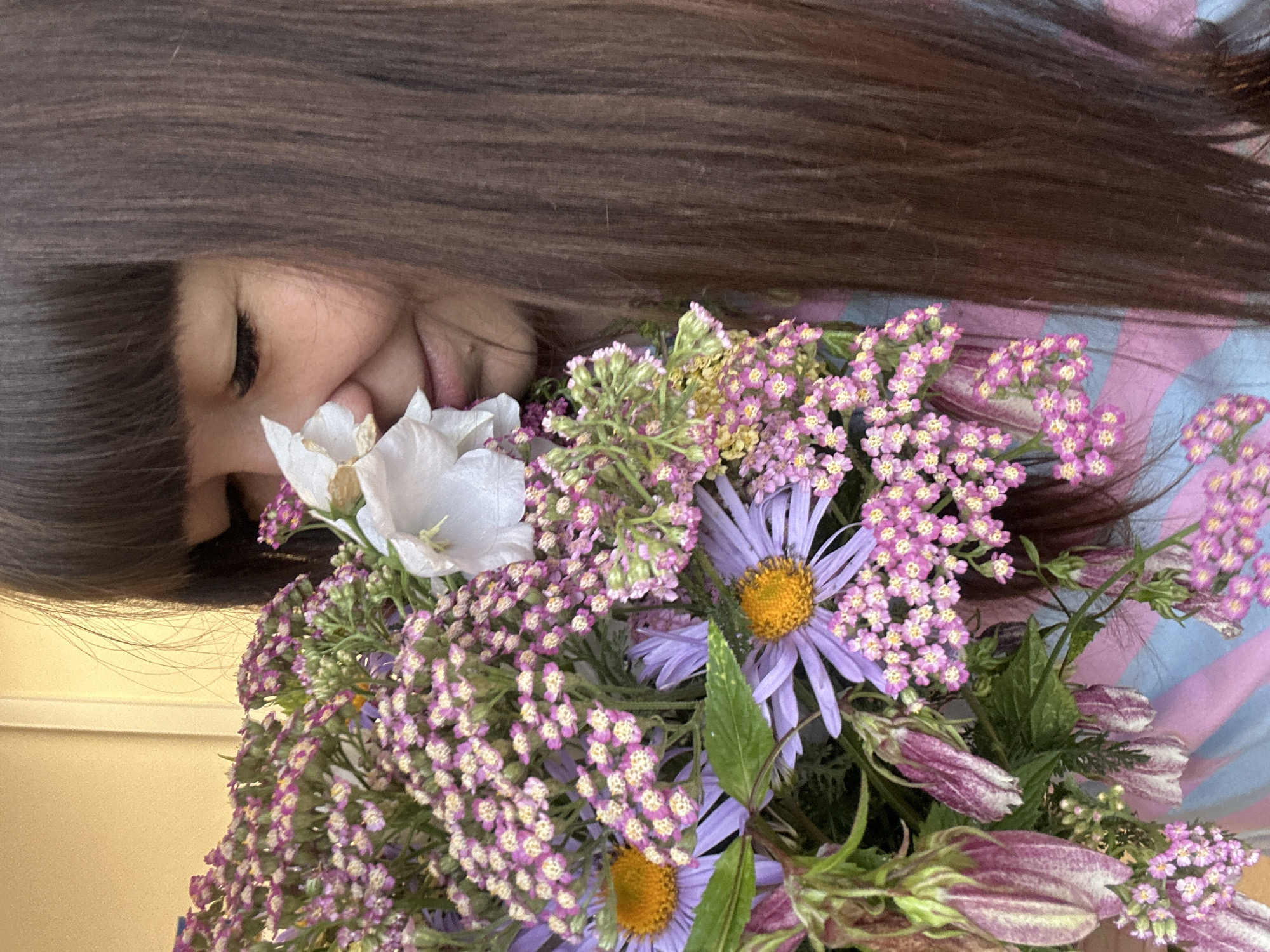 A young woman with long, dark brown hair and bangs is holding a bouquet of flowers, smelling one of the white flowers. The bouquet contains purple asters, small pink yarrow flowers, and some unopened bell-shaped flowers. She is wearing a blue and pink striped shirt. The background is a plain beige wall and a white vertical trim. The scene appears to be outdoors, possibly in a garden or on a porch, in the city of Chaikovsky, Russia.
