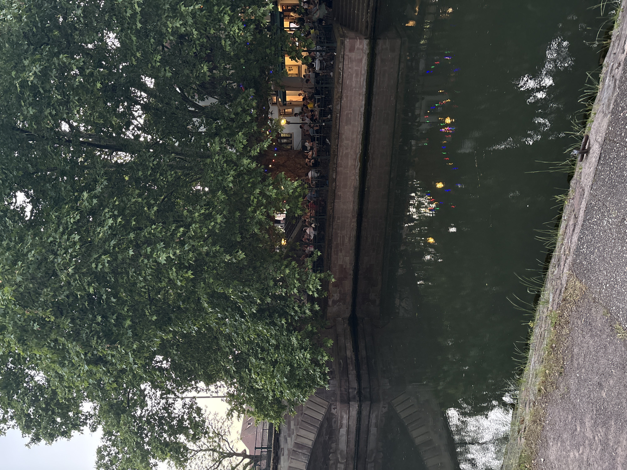The image shows a canal scene in Strasbourg, France. A tree occupies much of the left side of the frame. Along the canal edge is a restaurant or bar with numerous people seated at tables, illuminated by lights. Reflections of these lights are visible on the water's surface. A stone wall runs alongside the canal, with an arched bridge section visible in the lower left corner. On the right side of the frame is a paved walkway with some vegetation. The scene appears to be at dusk or night.