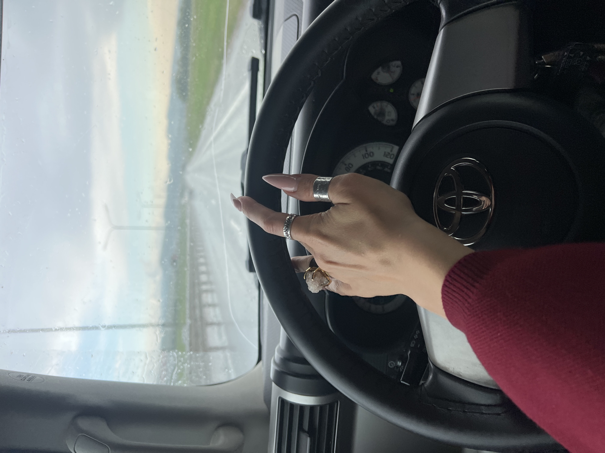 A person's hand with multiple rings is resting on the steering wheel of a Toyota vehicle. The vehicle appears to be driving on a wet road, as suggested by the droplets on the windshield and the visibility conditions. The surrounding environment is blurred, indicating motion. It is not possible to confirm the location in the city of Чистенское сельское поселение, Ukraine, from this image.