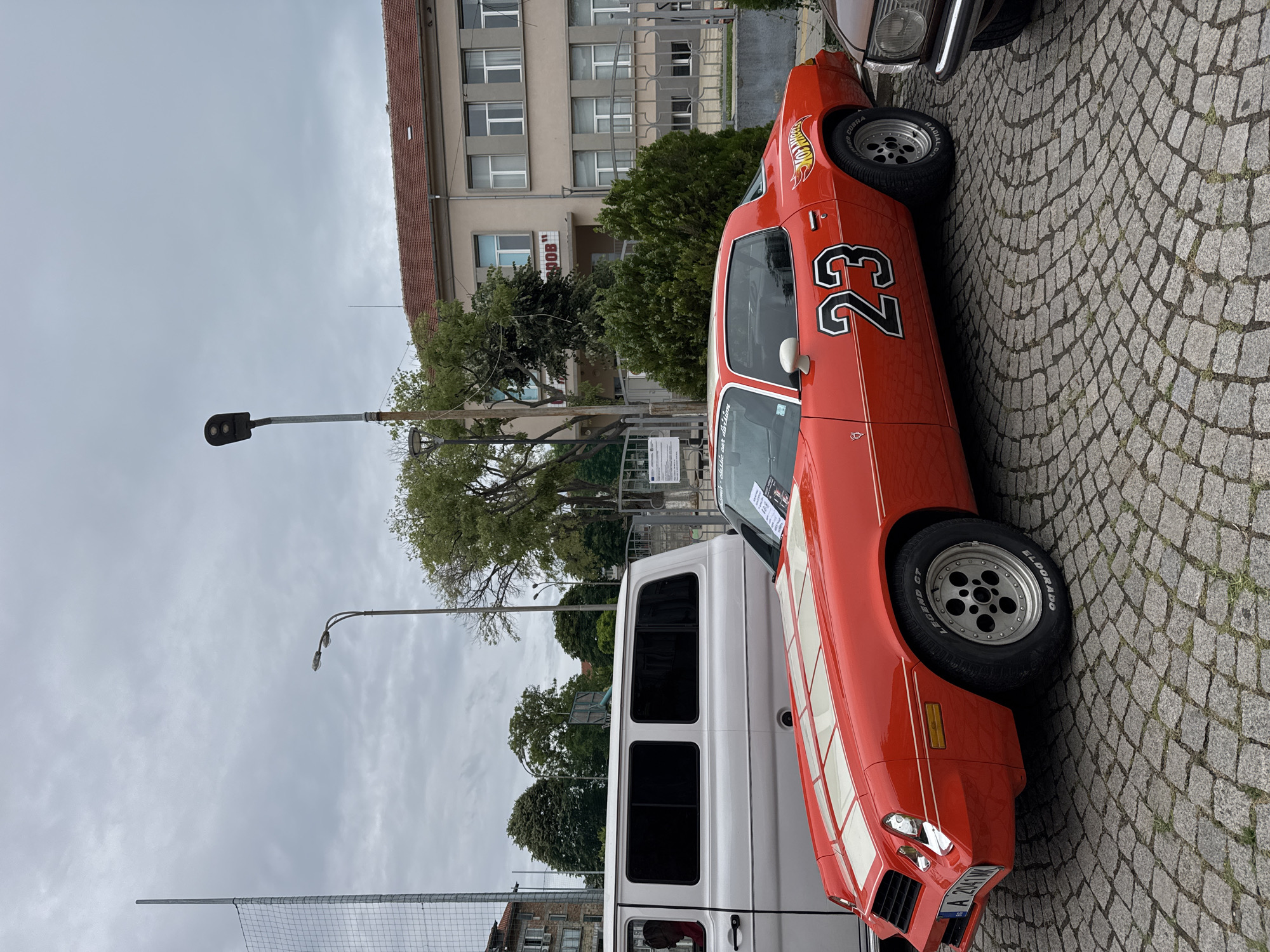 This is a medium shot, eye-level, outdoor photograph taken on a cloudy day. The foreground features a bright red Chevrolet Camaro with white racing stripes on the hood, displaying the number "23" prominently on its side, along with a "Hot Wheels" logo. The car has custom wheels and racing tires. To the left of the Camaro, a white van is partially visible. The ground is paved with cobblestones. In the background, there is a building with a tiled roof and multiple windows, and some trees and foliage. A street lamp is visible on the right side of the frame. The location is the main square in Tsarevo, Bulgaria.